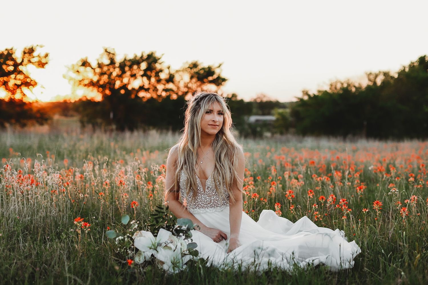 Bride in white dress sits among wildflowers during sunset in rustic field setting.