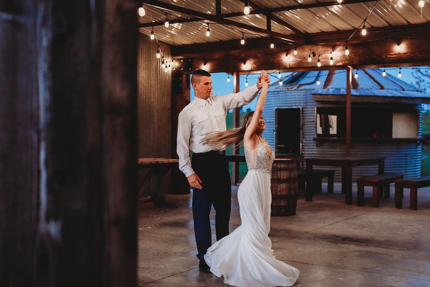 Couple dancing in rustic venue with string lights and wooden beams overhead.