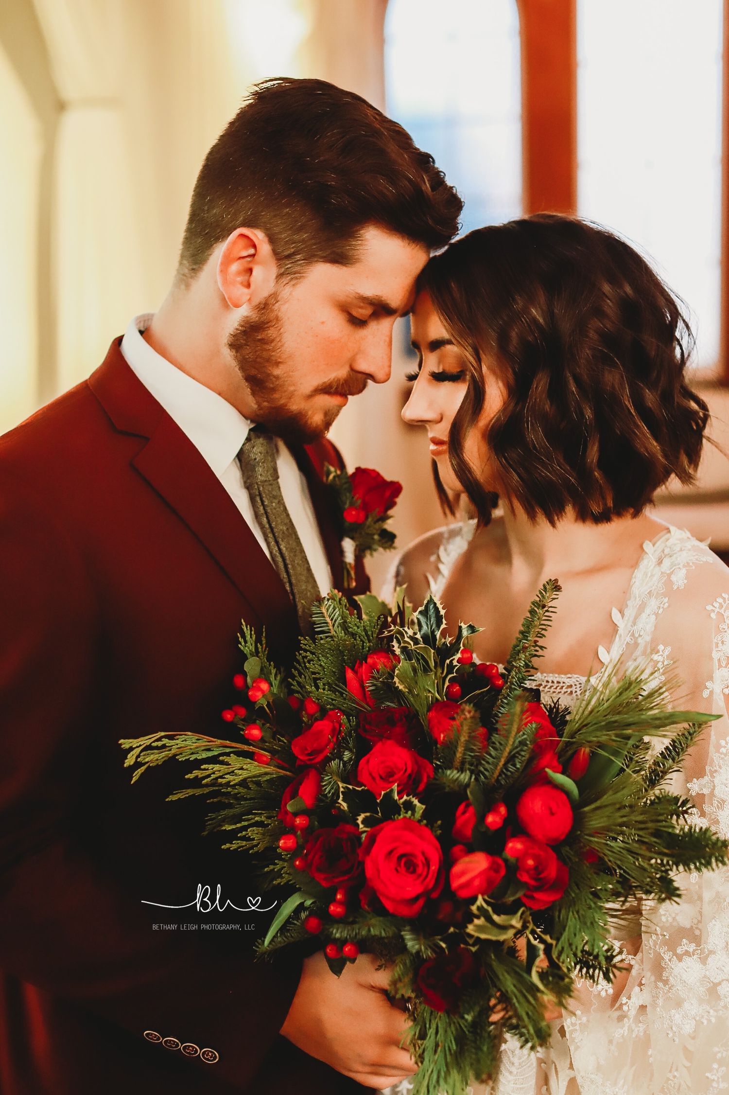 An intimate wedding portrait featuring a burgundy suit and winter bouquet with red roses.