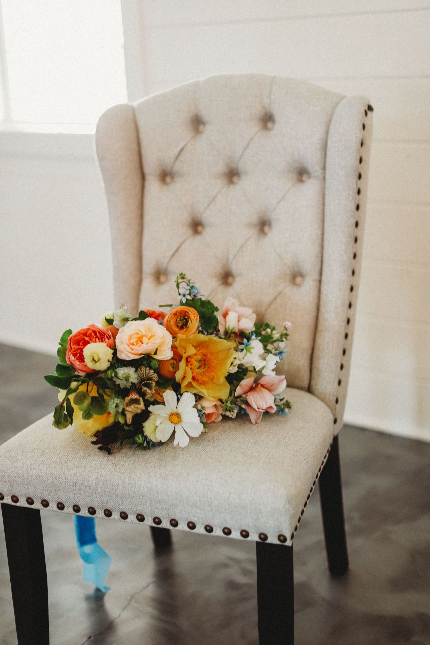 A vibrant floral bouquet with orange, pink and white flowers sits atop an elegant tufted beige dining chair with nailhead trim.