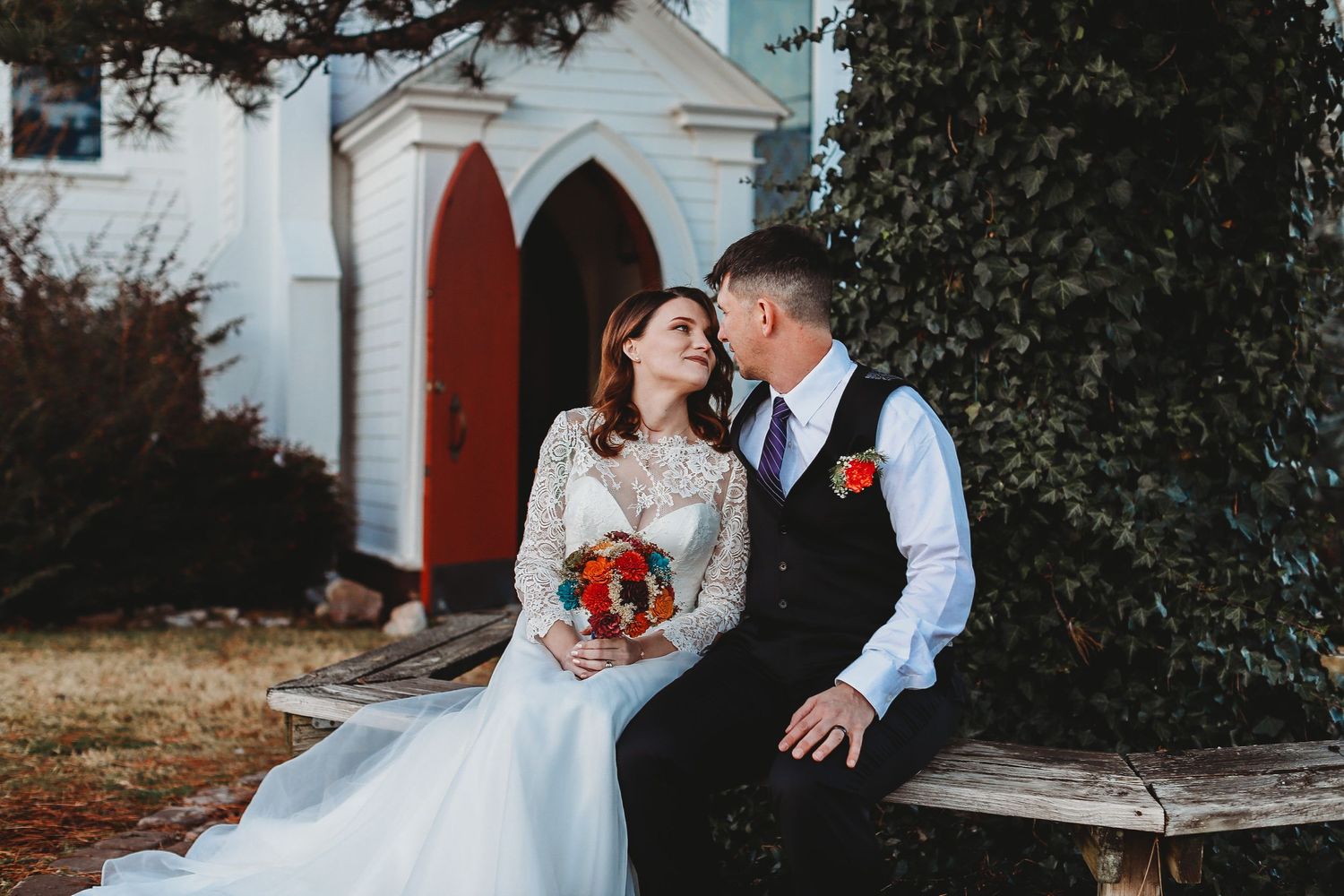 Couple in wedding attire sharing an intimate moment on a wooden bench outside a white church with Gothic arch.