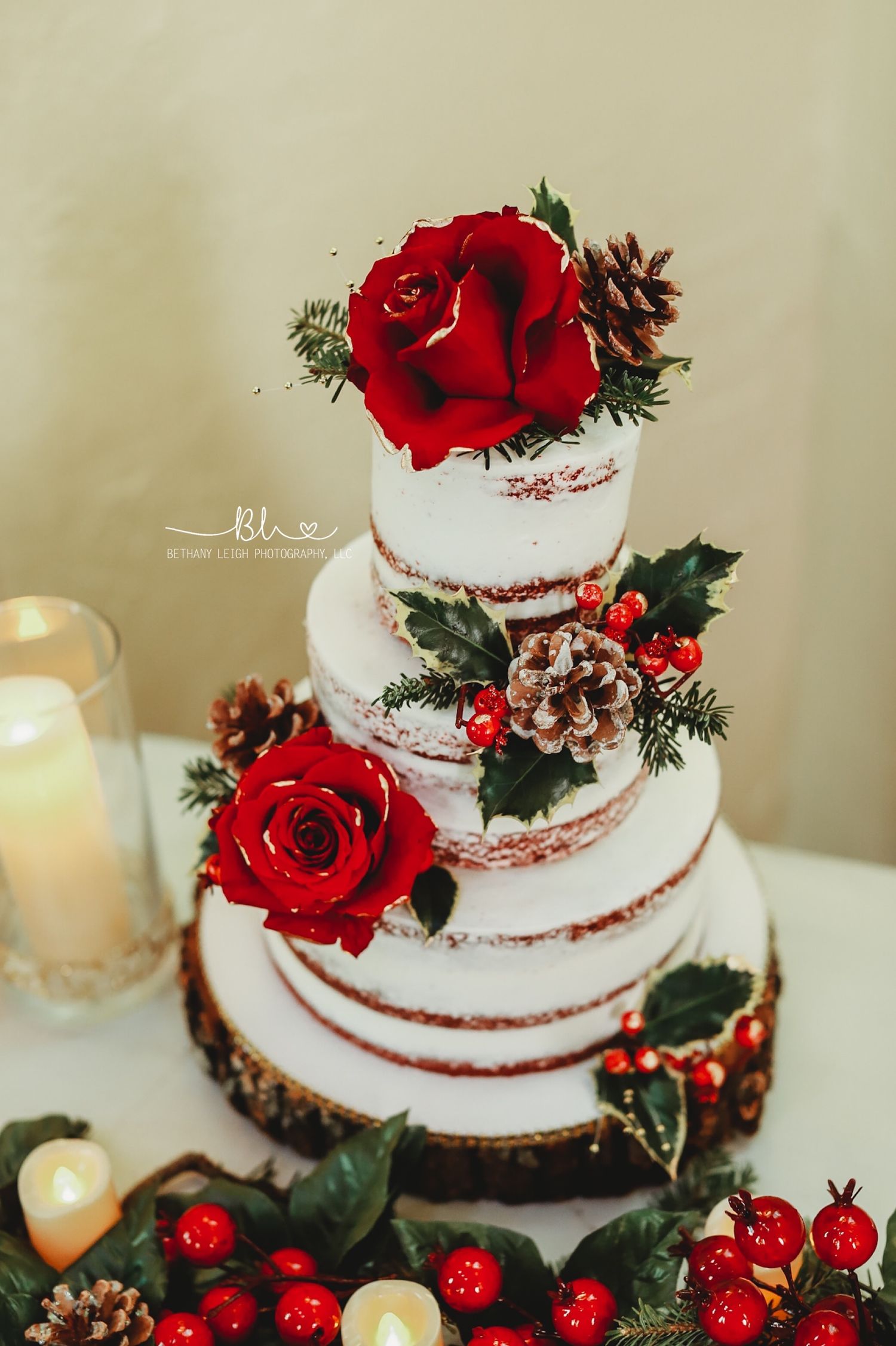 A semi-naked wedding cake decorated with red roses, holly berries, and pinecones.