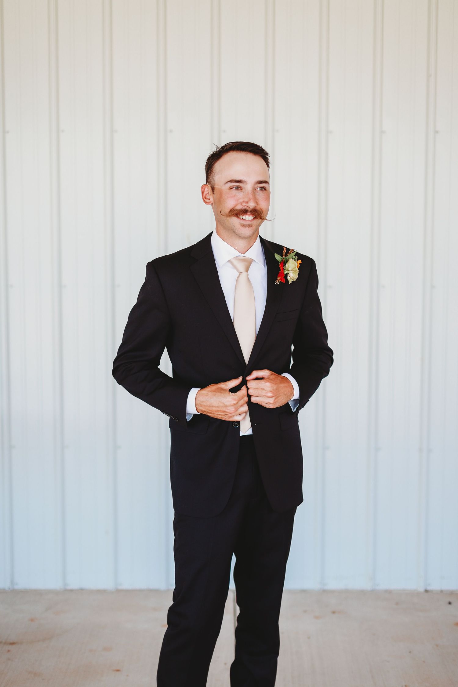 Groom in classic black tuxedo with peach tie and boutonniere against white backdrop.