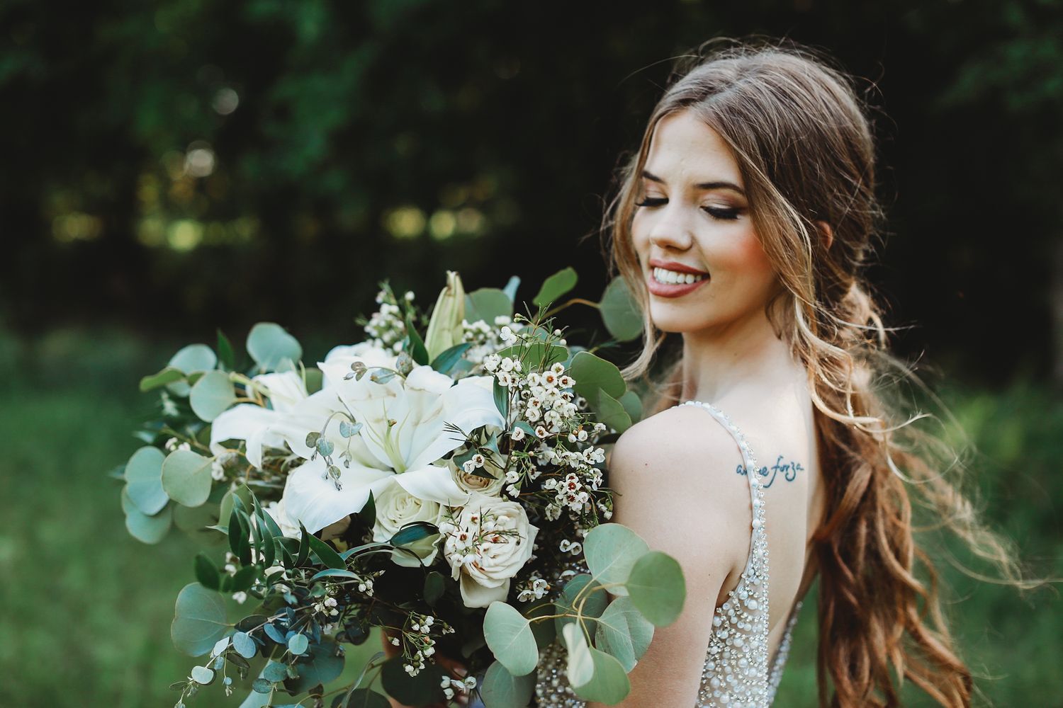 Bride with loose curls holds white and green bouquet while posing in outdoor garden setting.