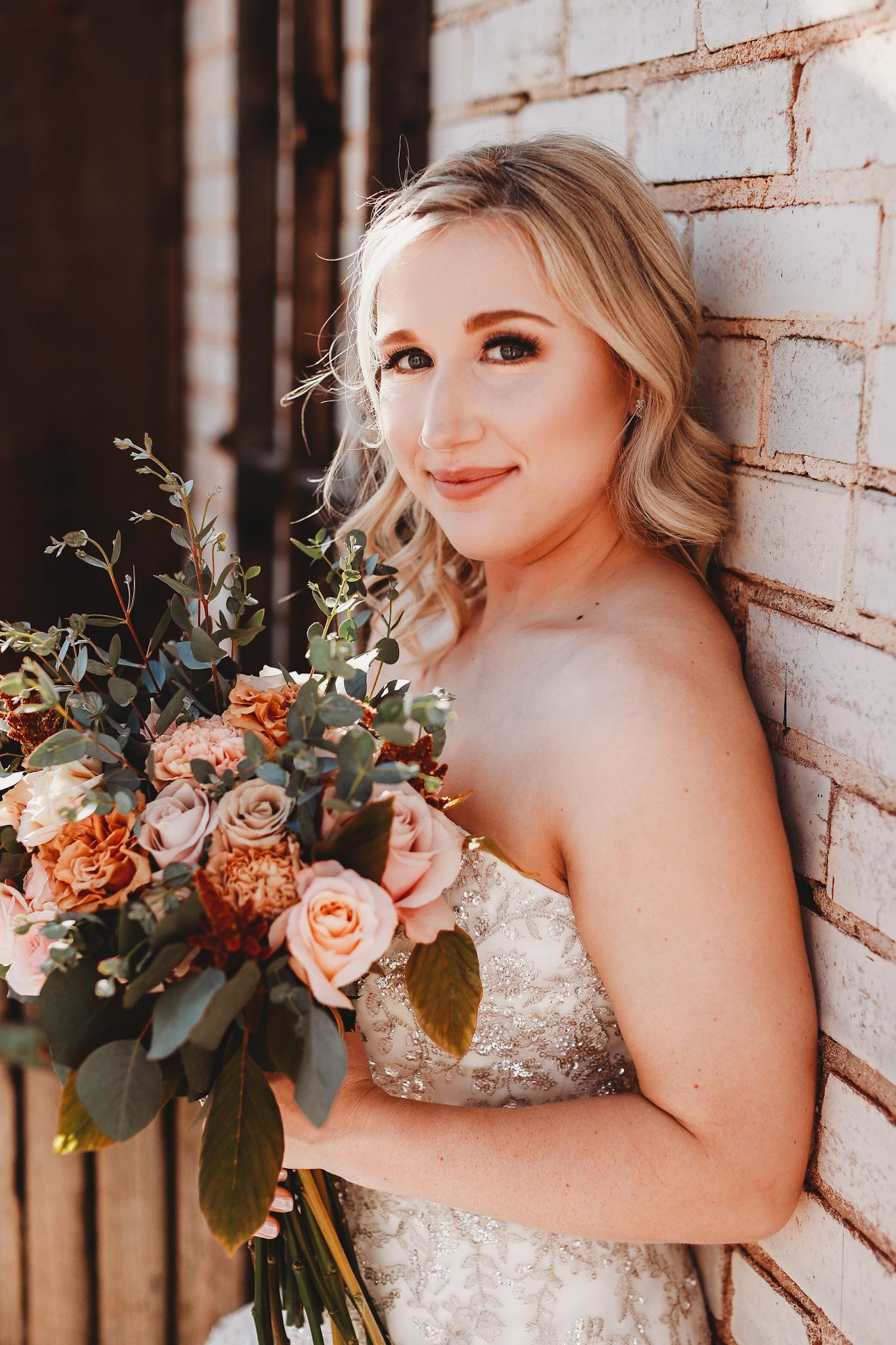 Bride with peach and blush floral bouquet poses against brick wall in strapless wedding gown.