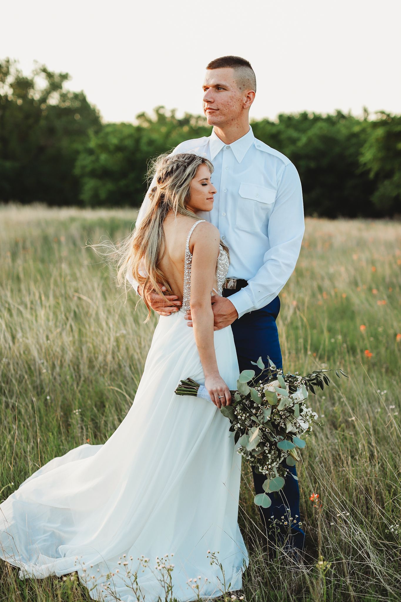 Couple embraces in field at sunset, bride in flowing dress holding eucalyptus bouquet.