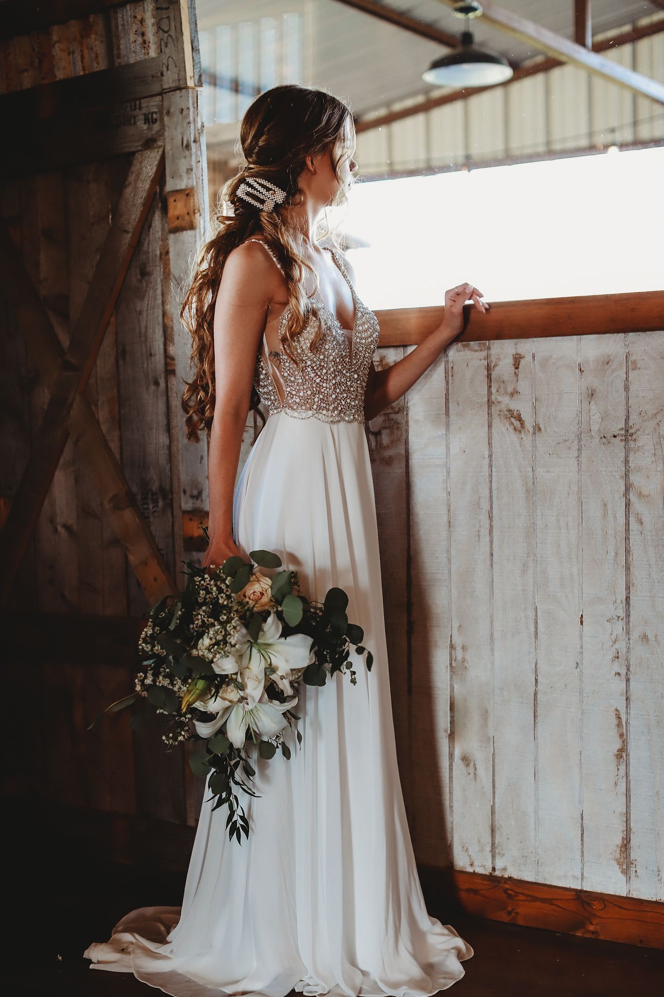 Bride in beaded white gown holds bouquet while standing against rustic wooden wall.