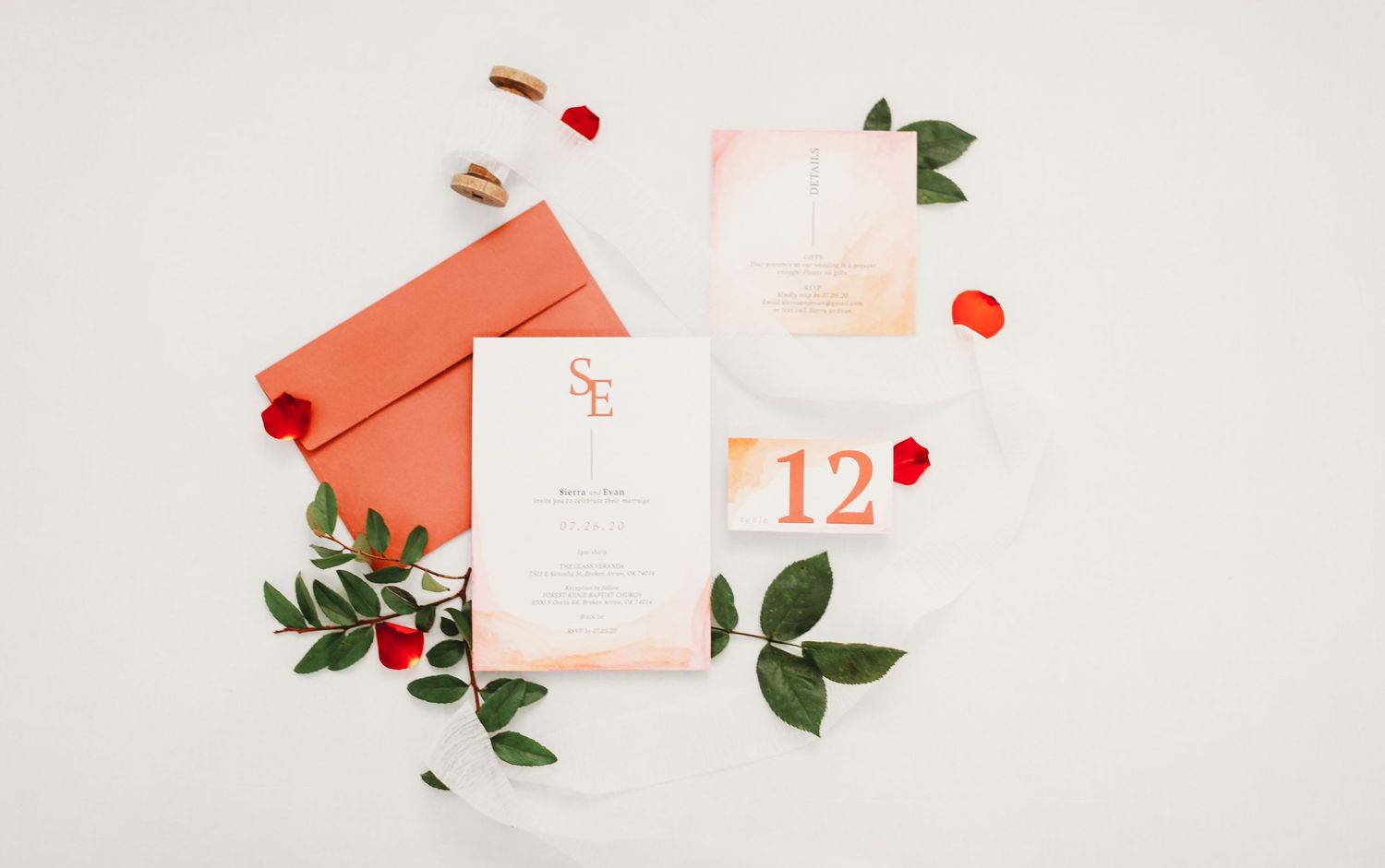 Wedding invitation suite with coral envelope, white stationery and delicate greenery on light beige background.