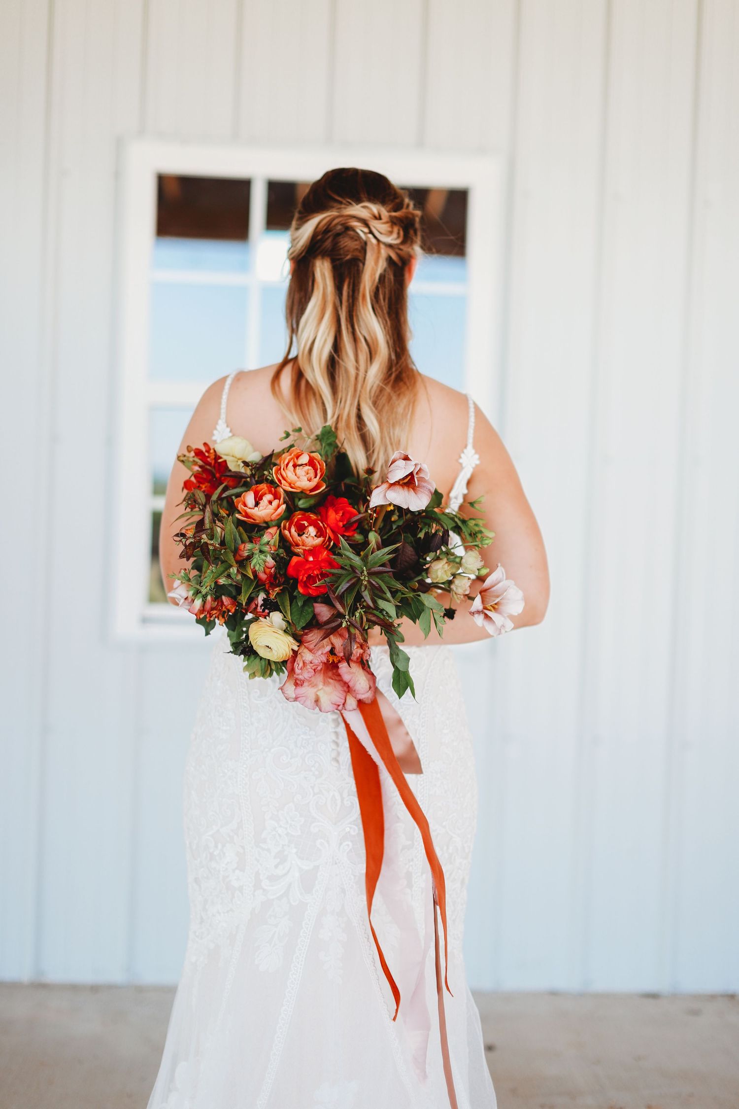Back view of bride in white lace gown holding vibrant red and pink floral bouquet with trailing red ribbons.