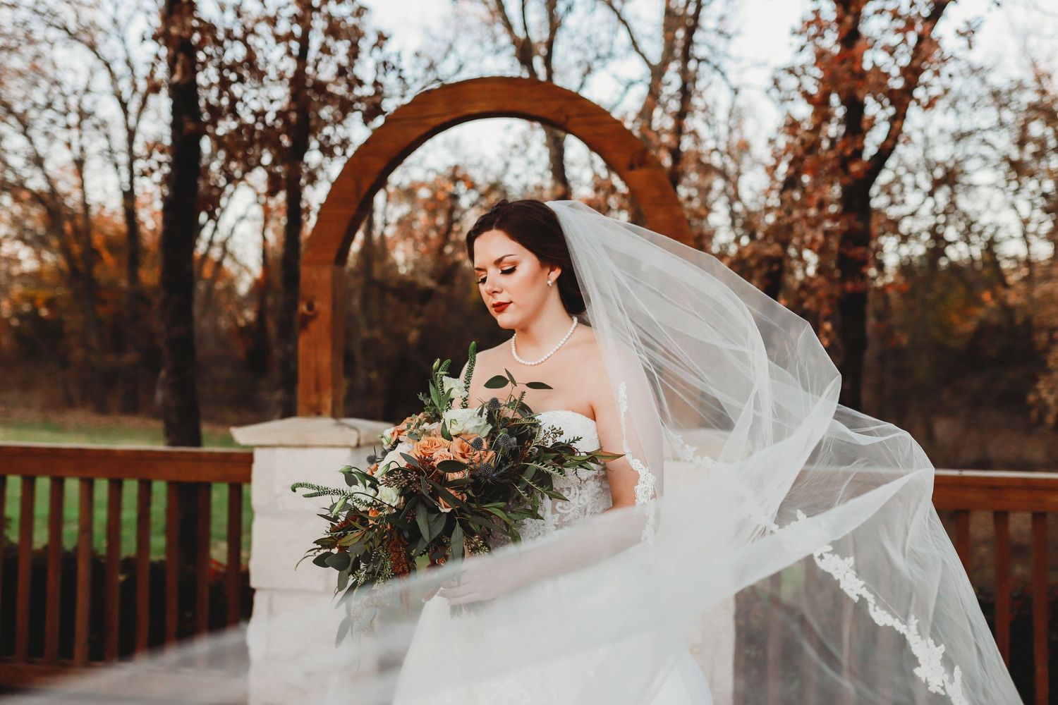 A dramatic bridal portrait with flowing veil and bouquet against a rustic wooden arch at sunset.