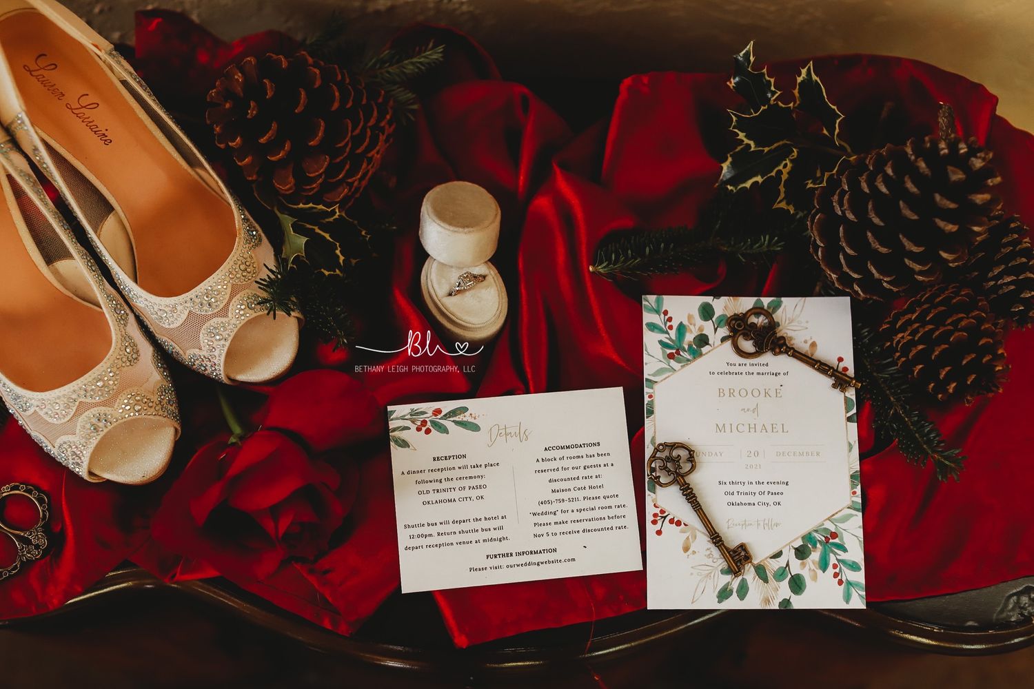 Wedding details flatlay with lace shoes, invitation, vintage key and pinecones on red fabric.