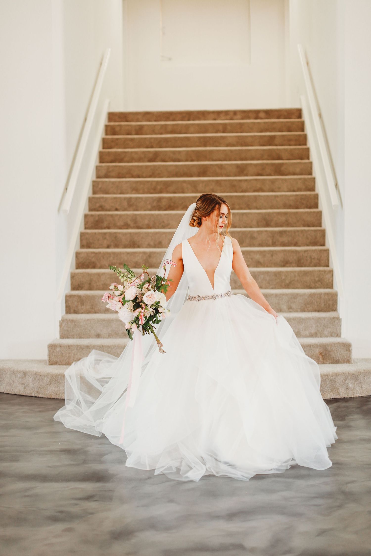 Bride in flowing ballgown poses on grand staircase holding floral bouquet.