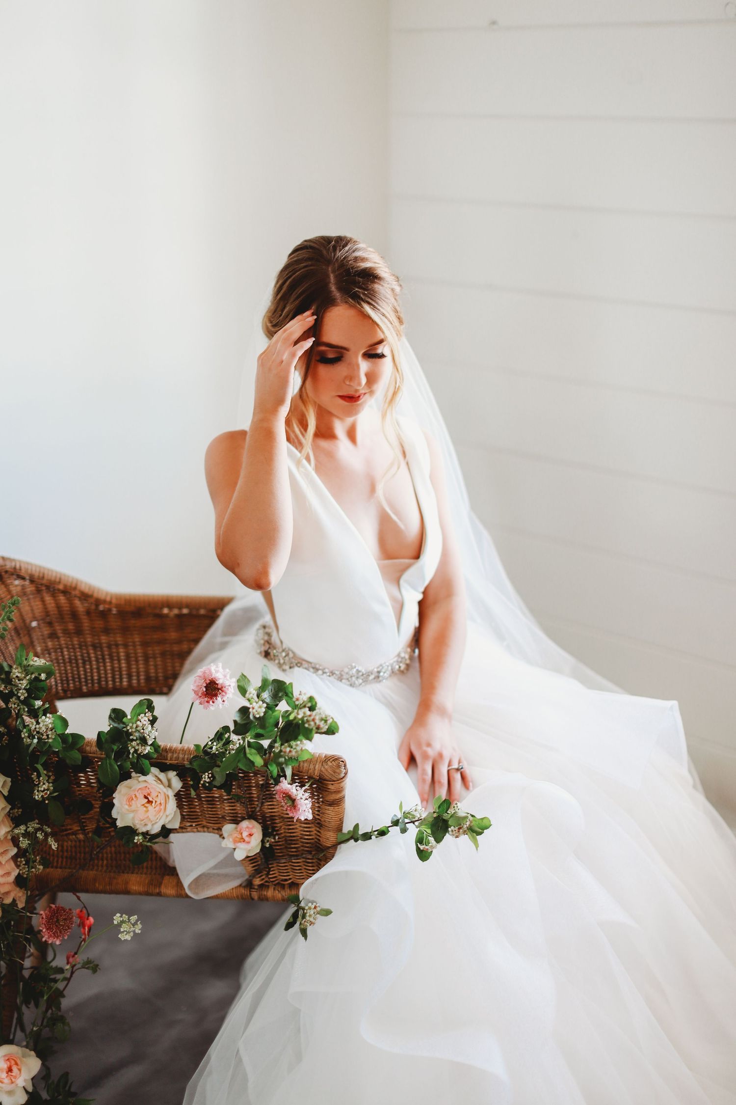 Bride in white gown sits thoughtfully by floral arrangements in rustic setting.