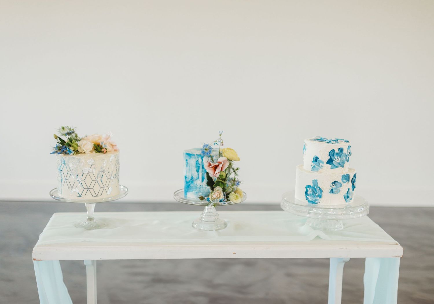 Three elegant white and blue wedding cakes displayed on a whitewashed table against a neutral background.