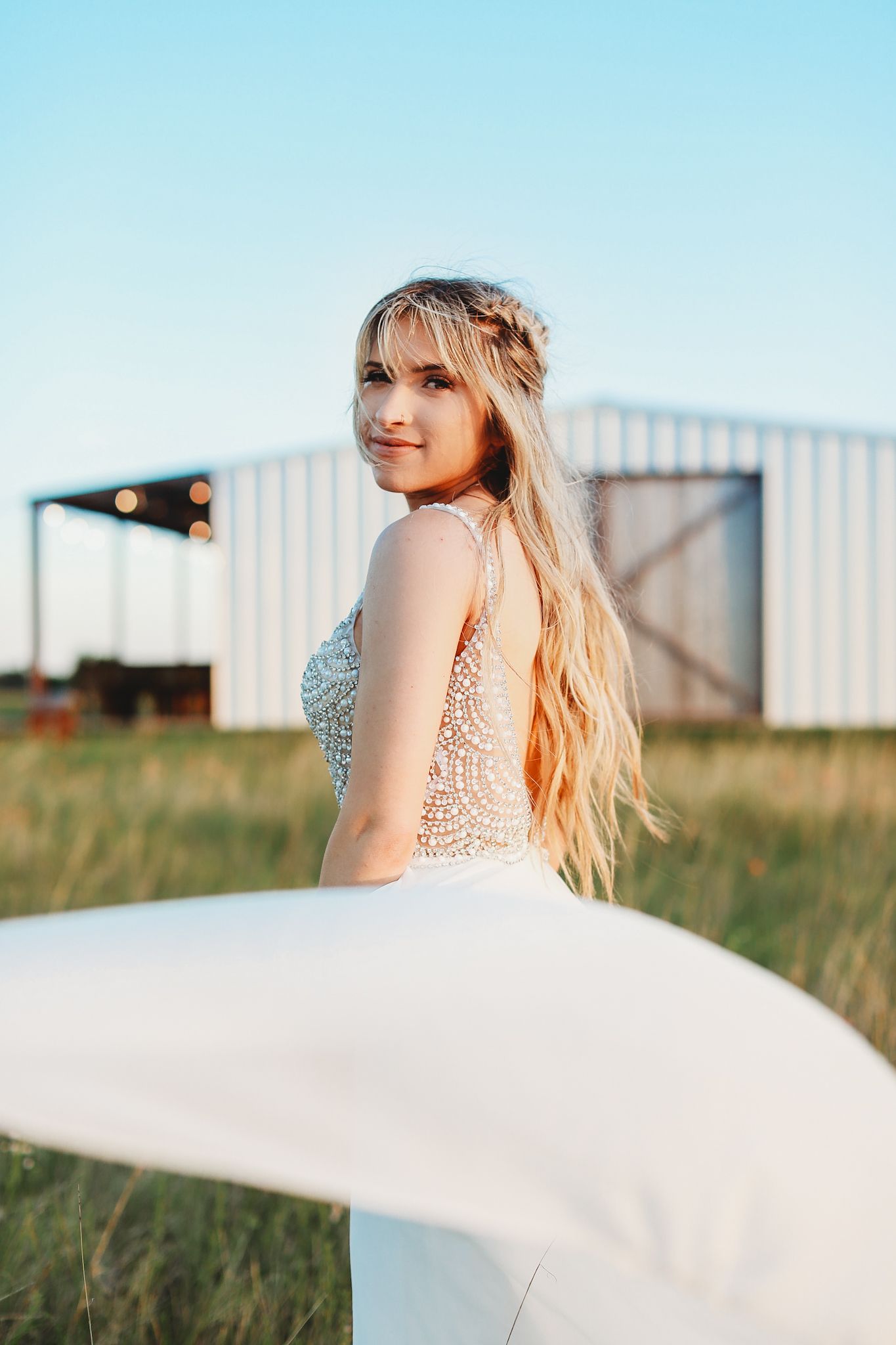 Bride in flowing white dress twirls in a field at golden hour with barn in background.