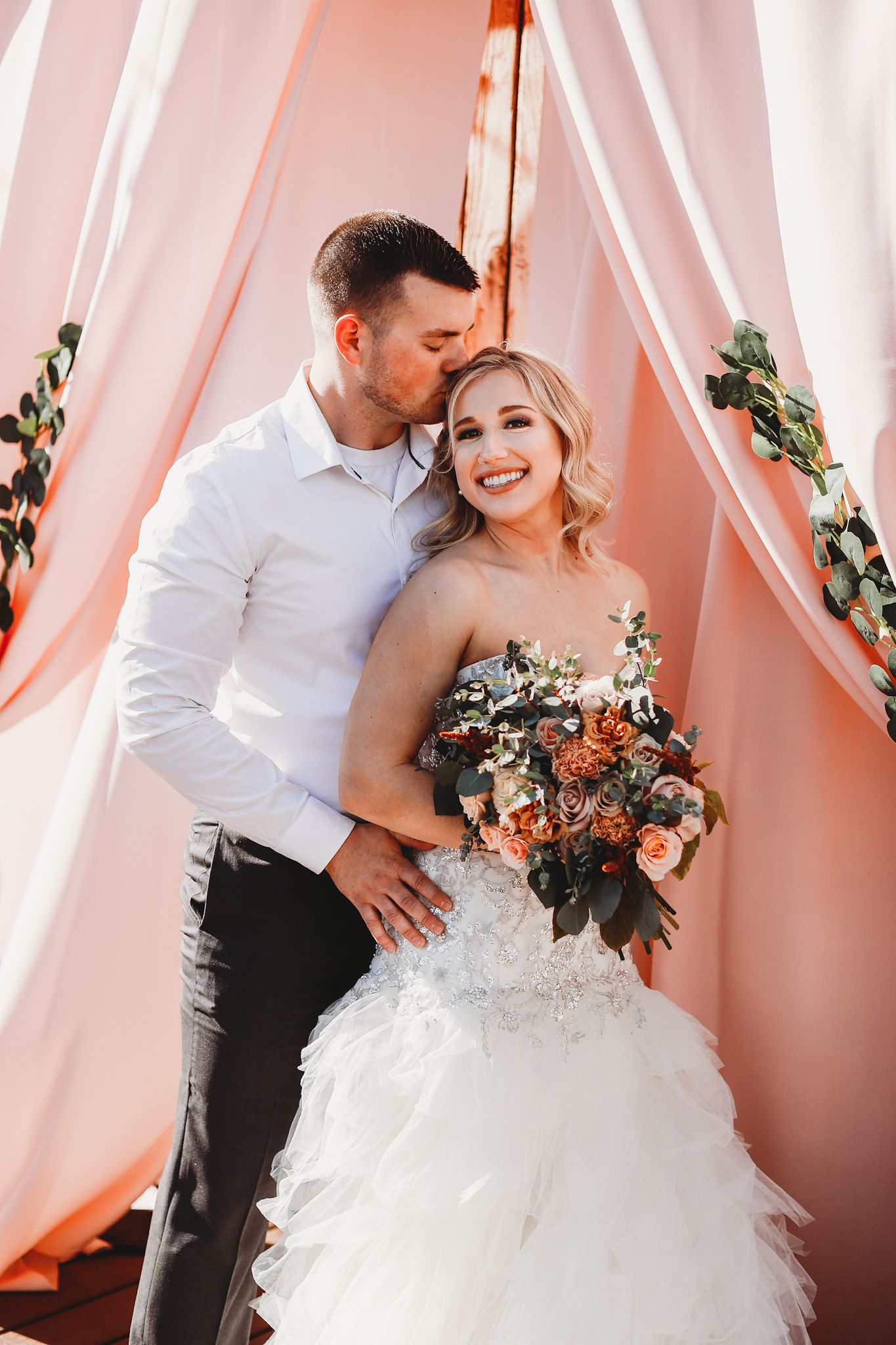 Wedding couple embraces under a pink draped arch decorated with floral arrangements and greenery.