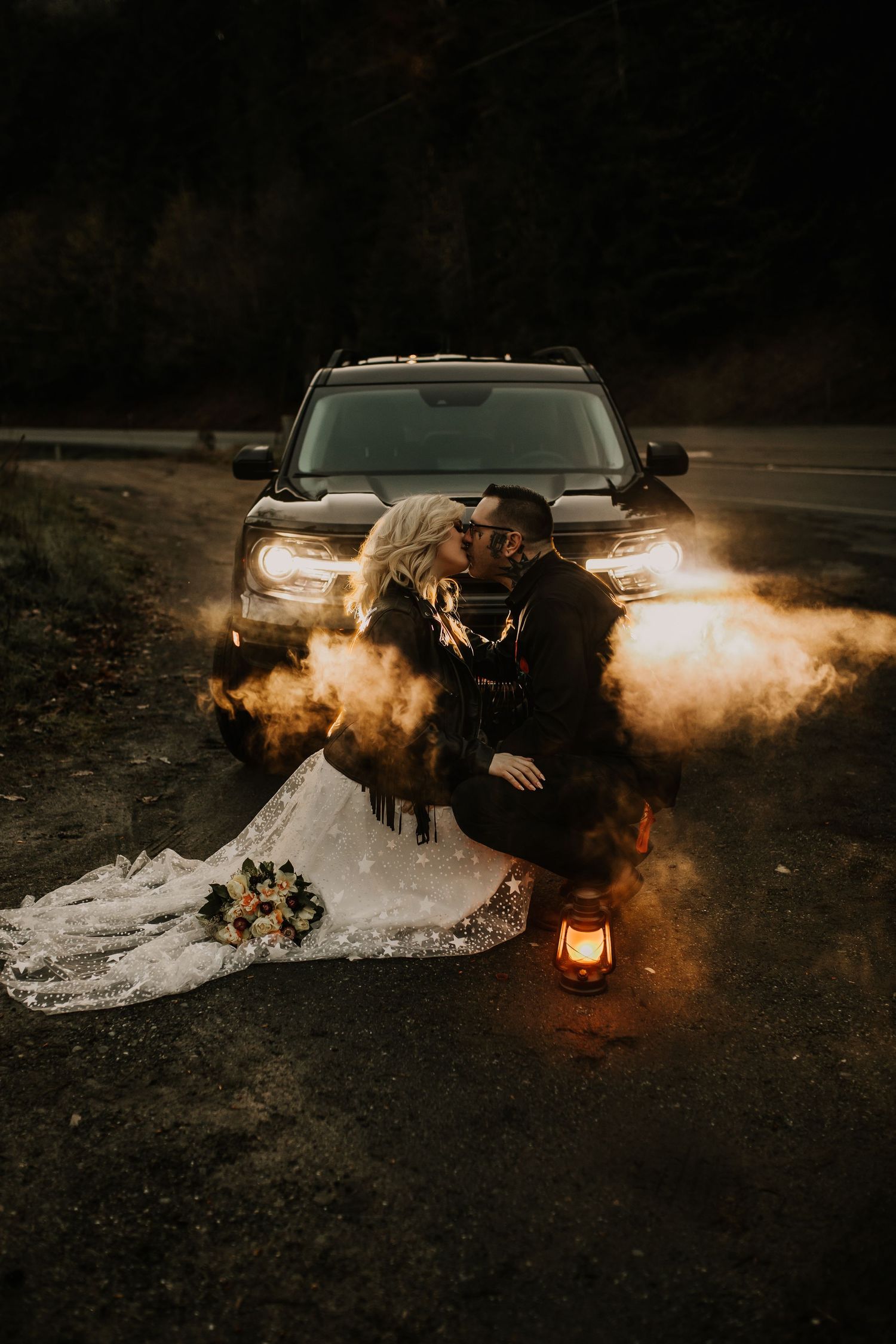 Bride and groom kissing in front of car headlights with glowing smoke and lantern at night.