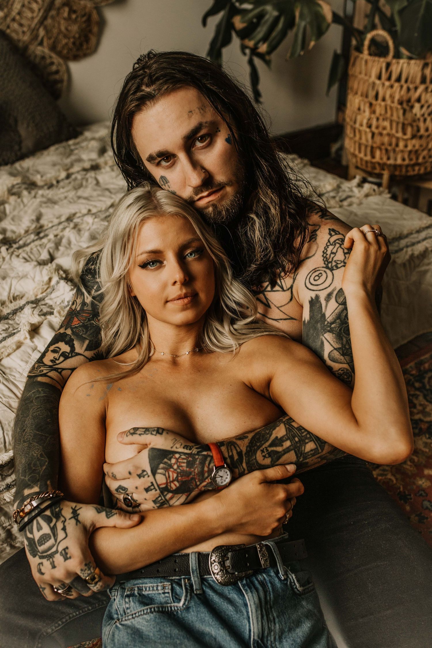 Heavily tattooed couple posing intimately in a bohemian bedroom, woman in jeans, man embracing her.