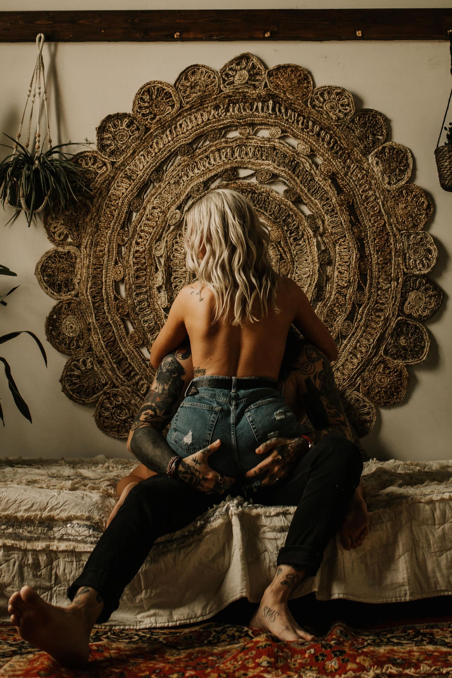 Tattooed woman sitting on a bed in front of a large ornate woven mandala wall hanging.