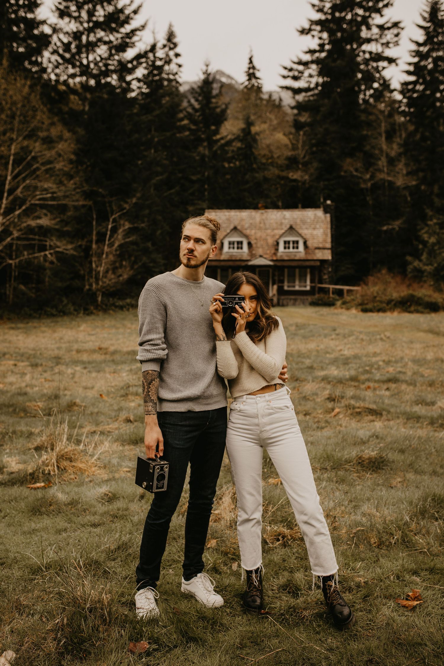 Couple poses in field with vintage camera before rustic cabin in forest.
