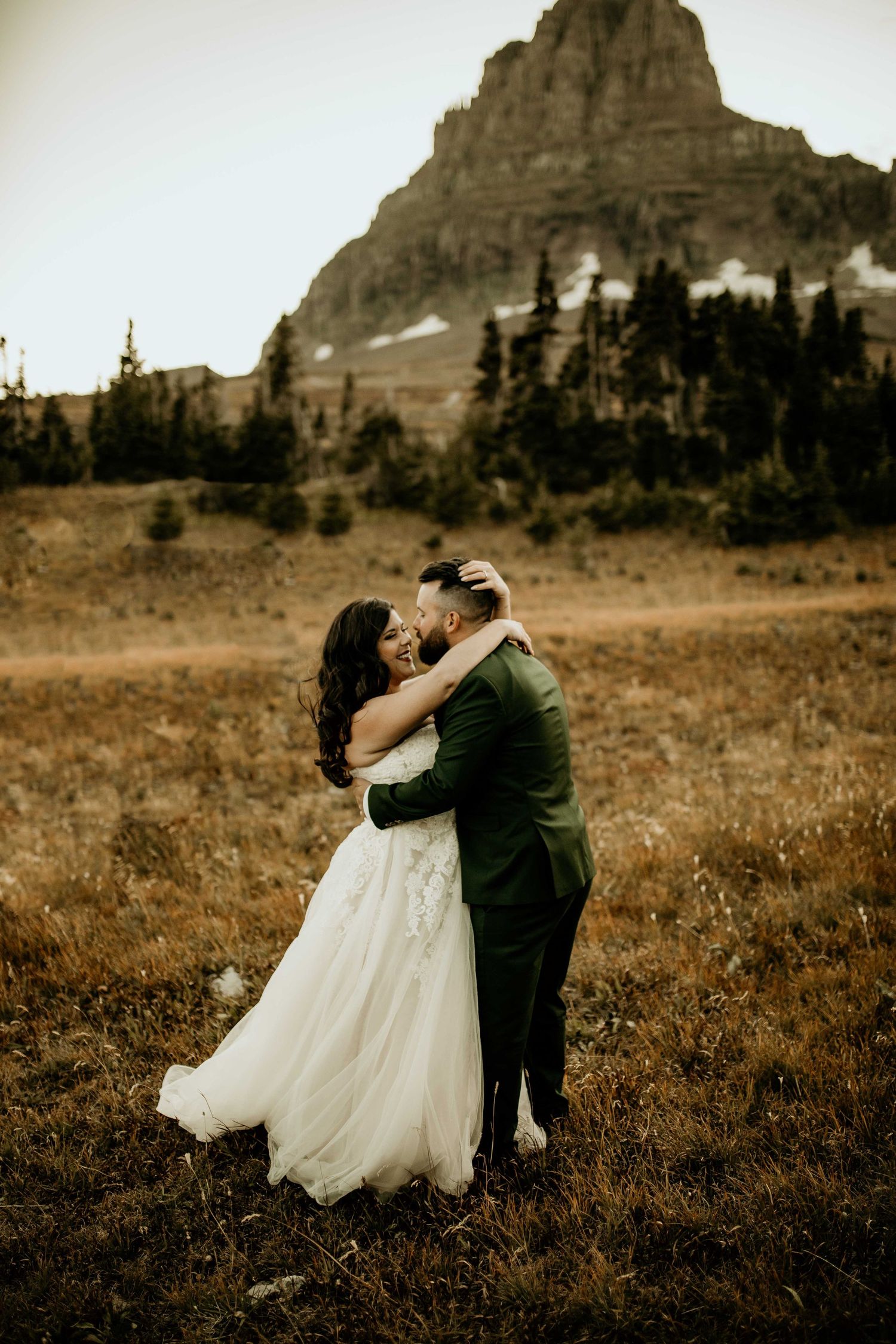 Bride and groom embracing in a mountain meadow with a rocky peak in the background.