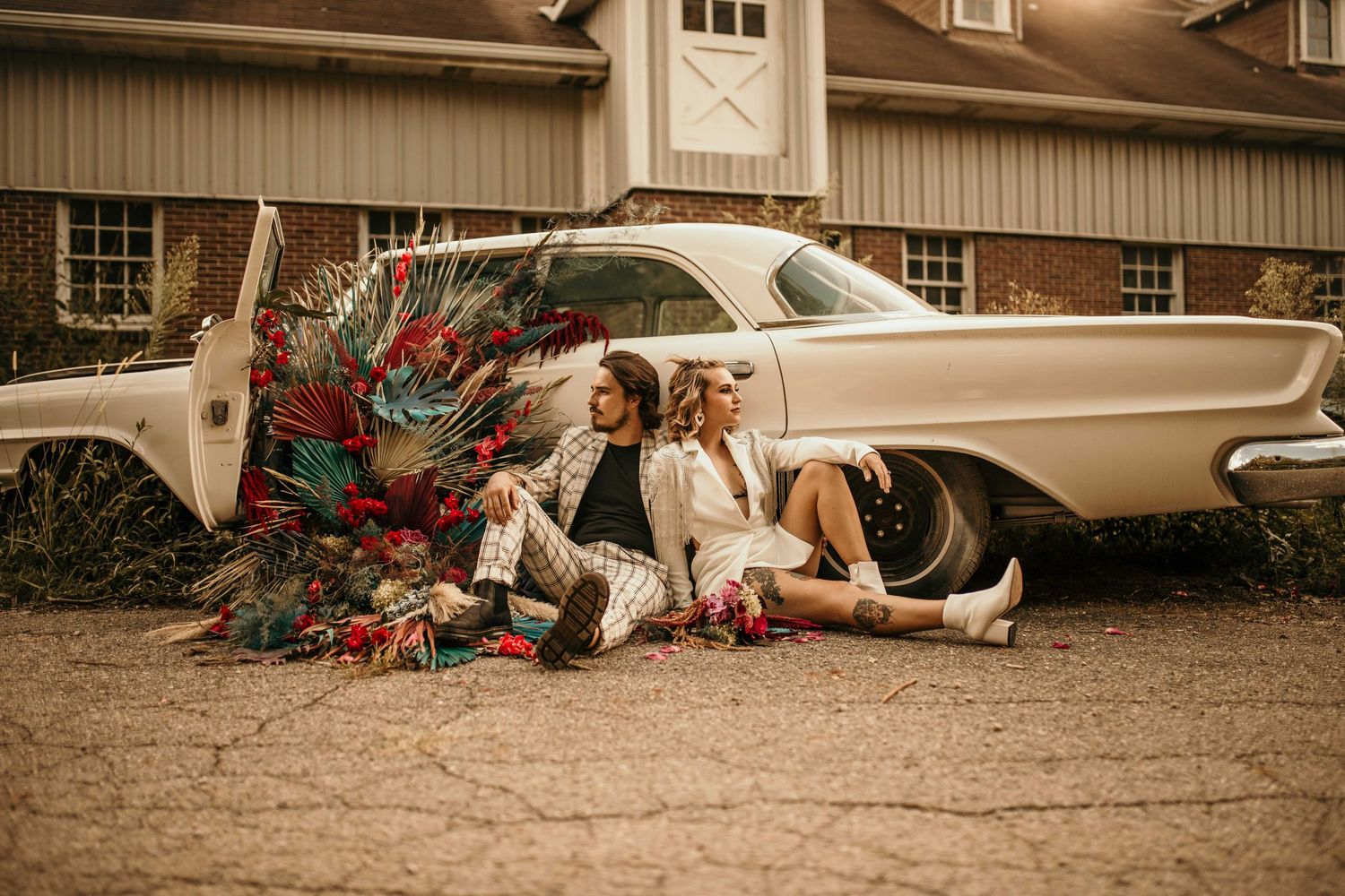 Stylish couple sitting against a vintage cream car with colorful tropical floral arrangement.