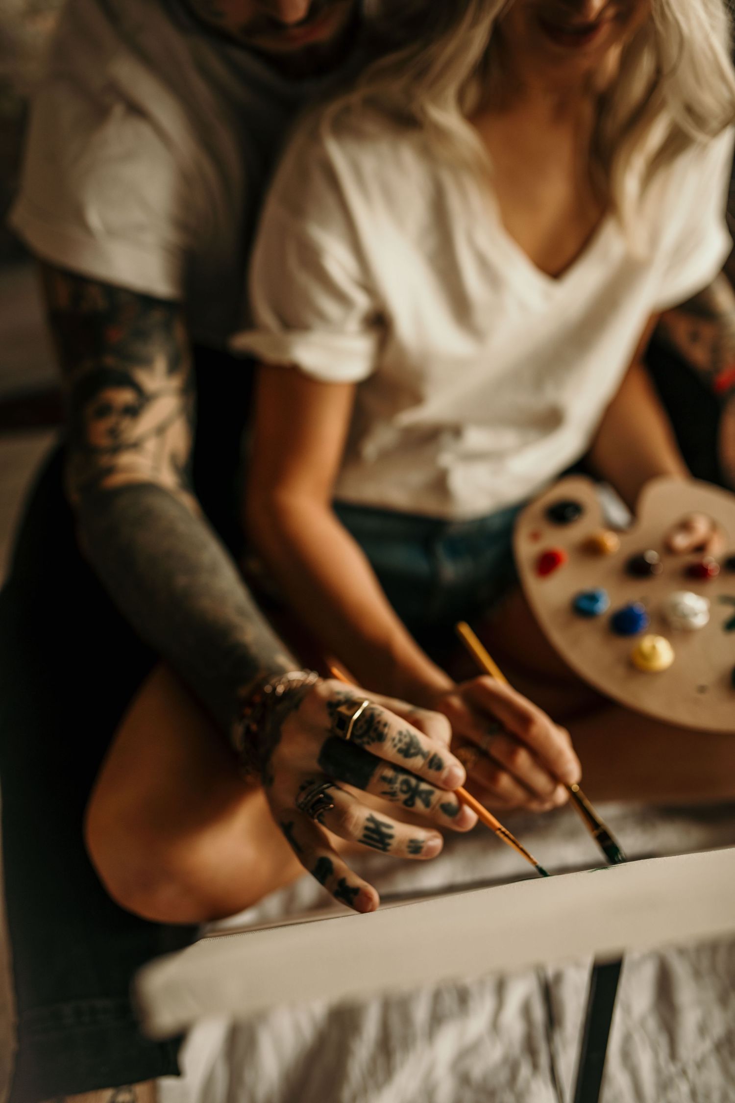 Tattooed couple sharing a creative moment painting together, holding brushes over a canvas.