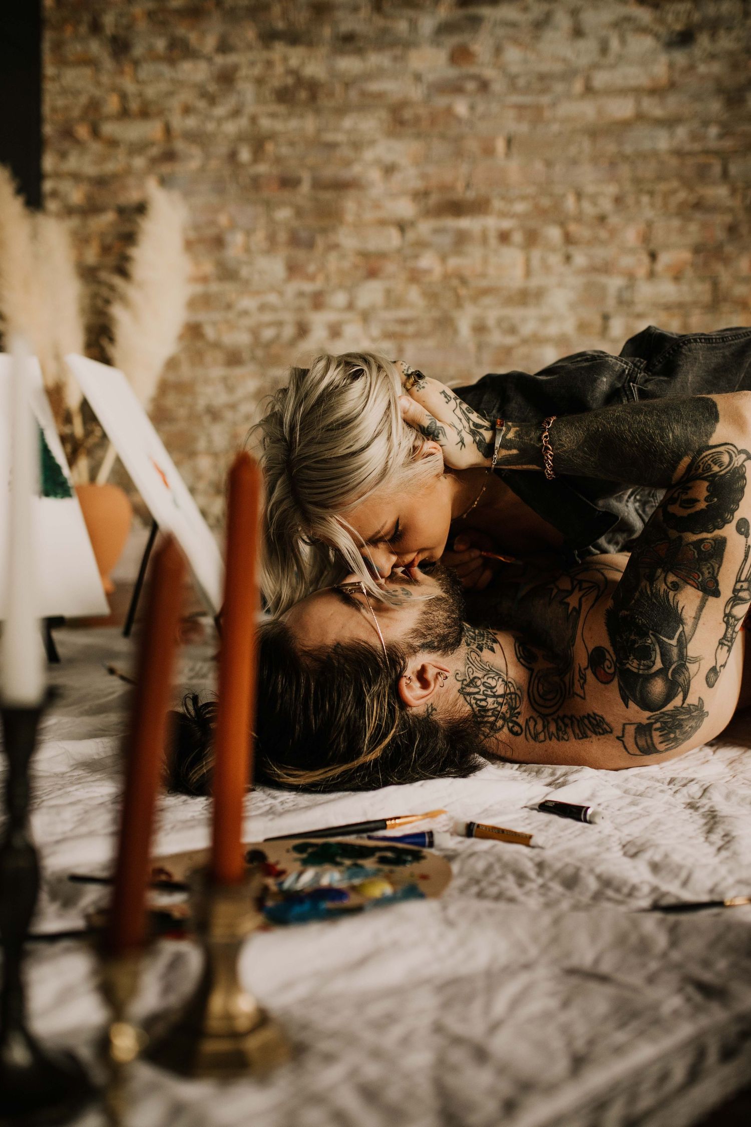 Tattooed couple sharing a kiss on a bed surrounded by candles and art supplies.