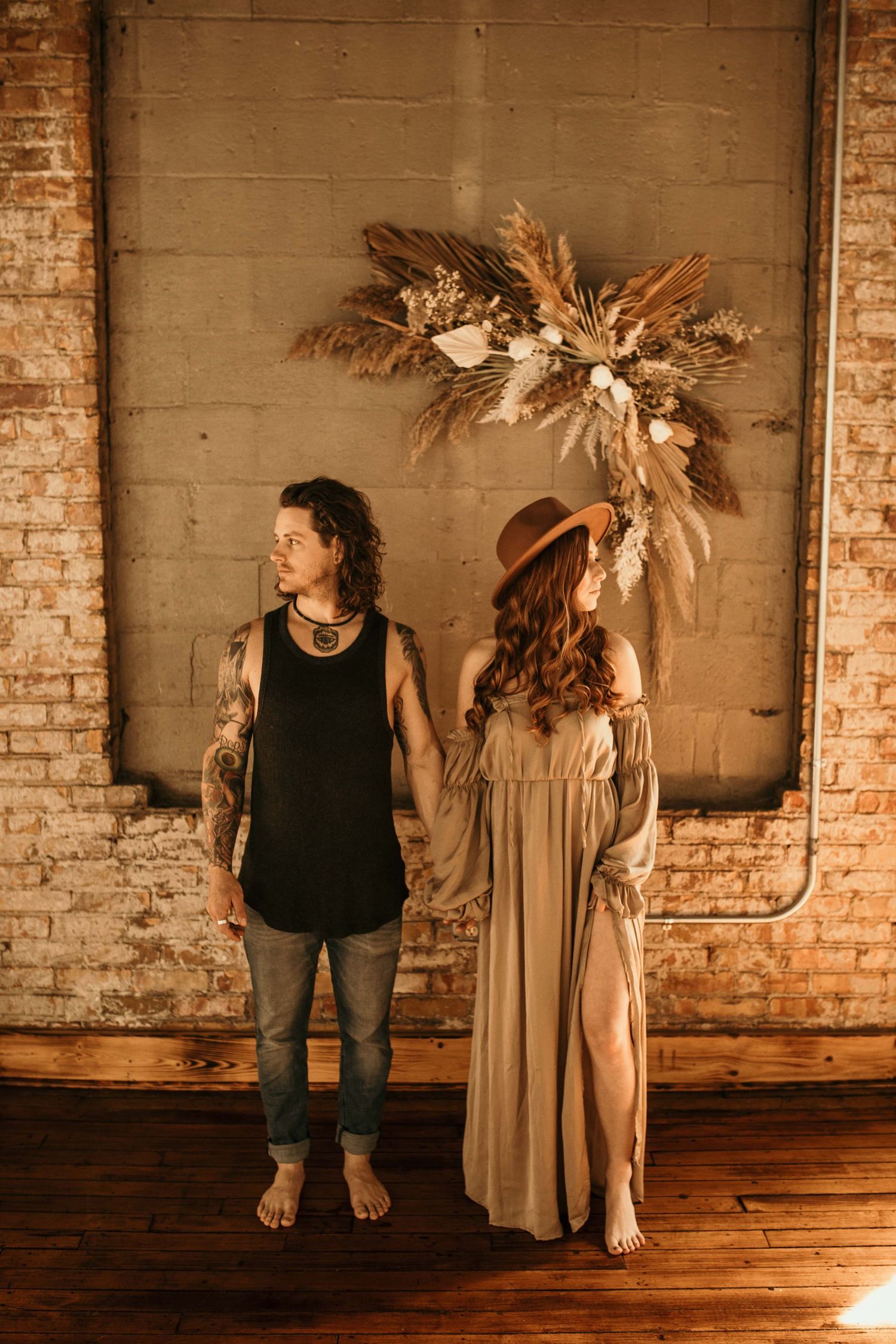 Bohemian couple holding hands in front of a dried floral arrangement on a brick wall.