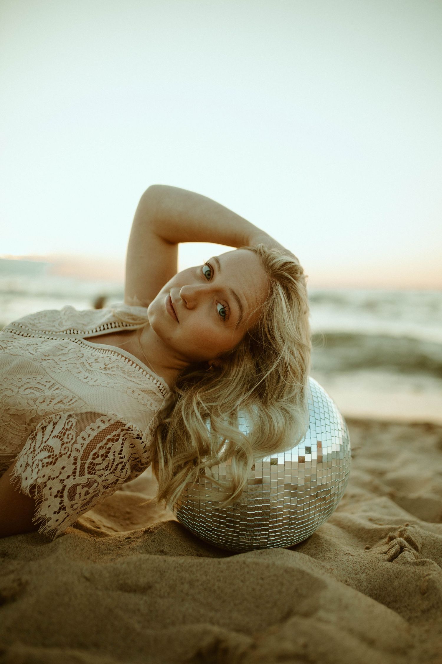 Blonde woman in lace top resting on disco ball on sandy beach at sunset.
