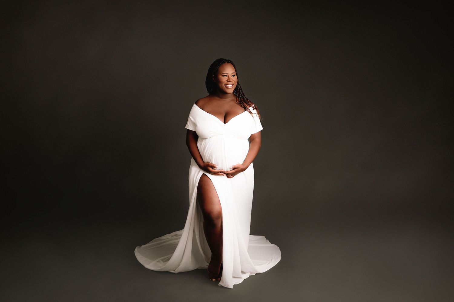 pregnant woman of color wearing white maternity dress on dark grey backdrop during professional studio maternity photos