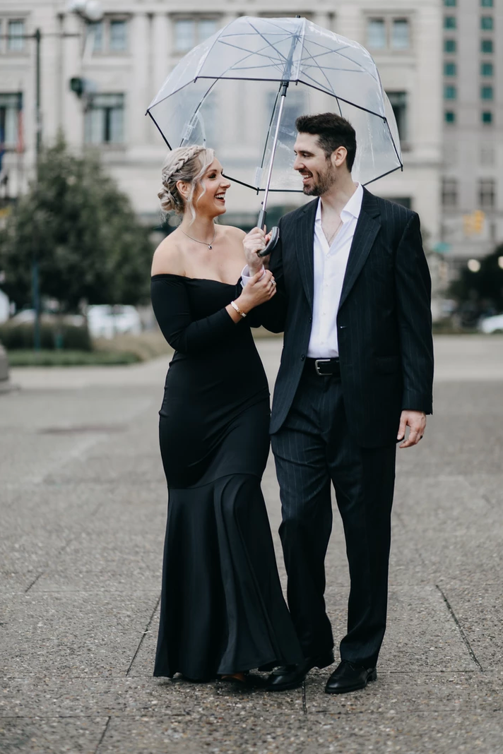Stylish couple share umbrella while walking in formal wear.