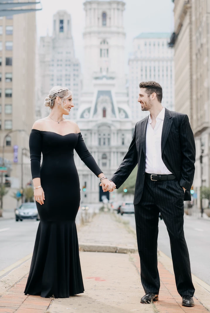 Couple in formal wear hold hands with historic architecture in background.