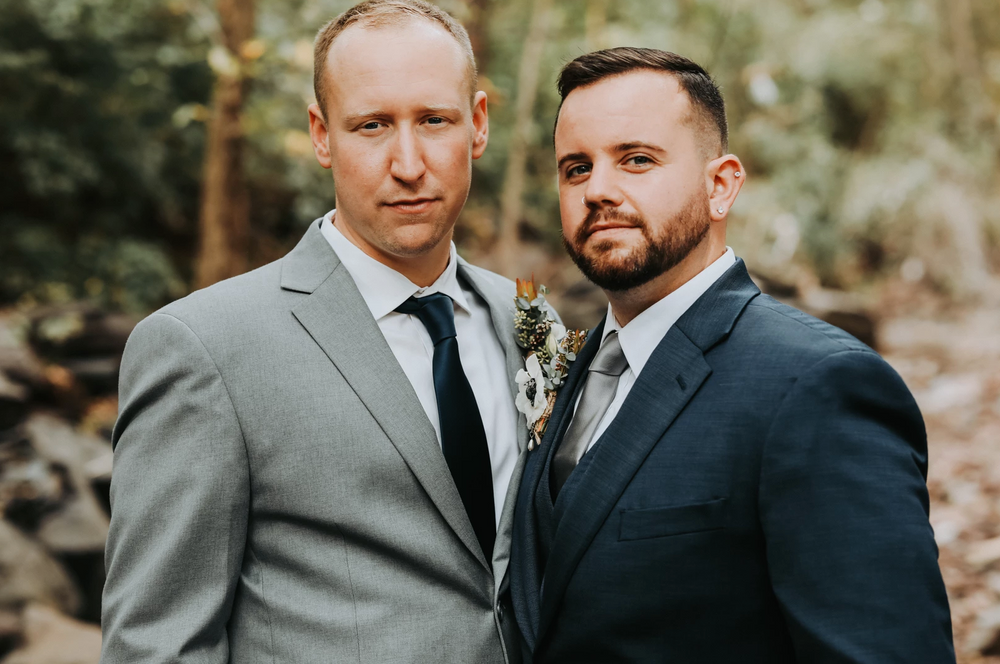 Two grooms in formal suits pose together outdoors for a wedding portrait.