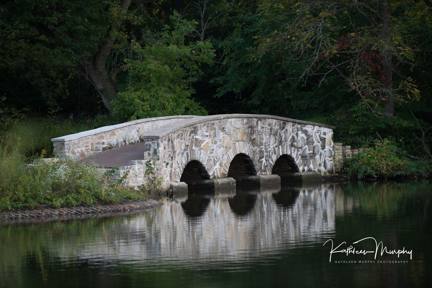 A stone bridge with three arches reflects perfectly in still water surrounded by trees.