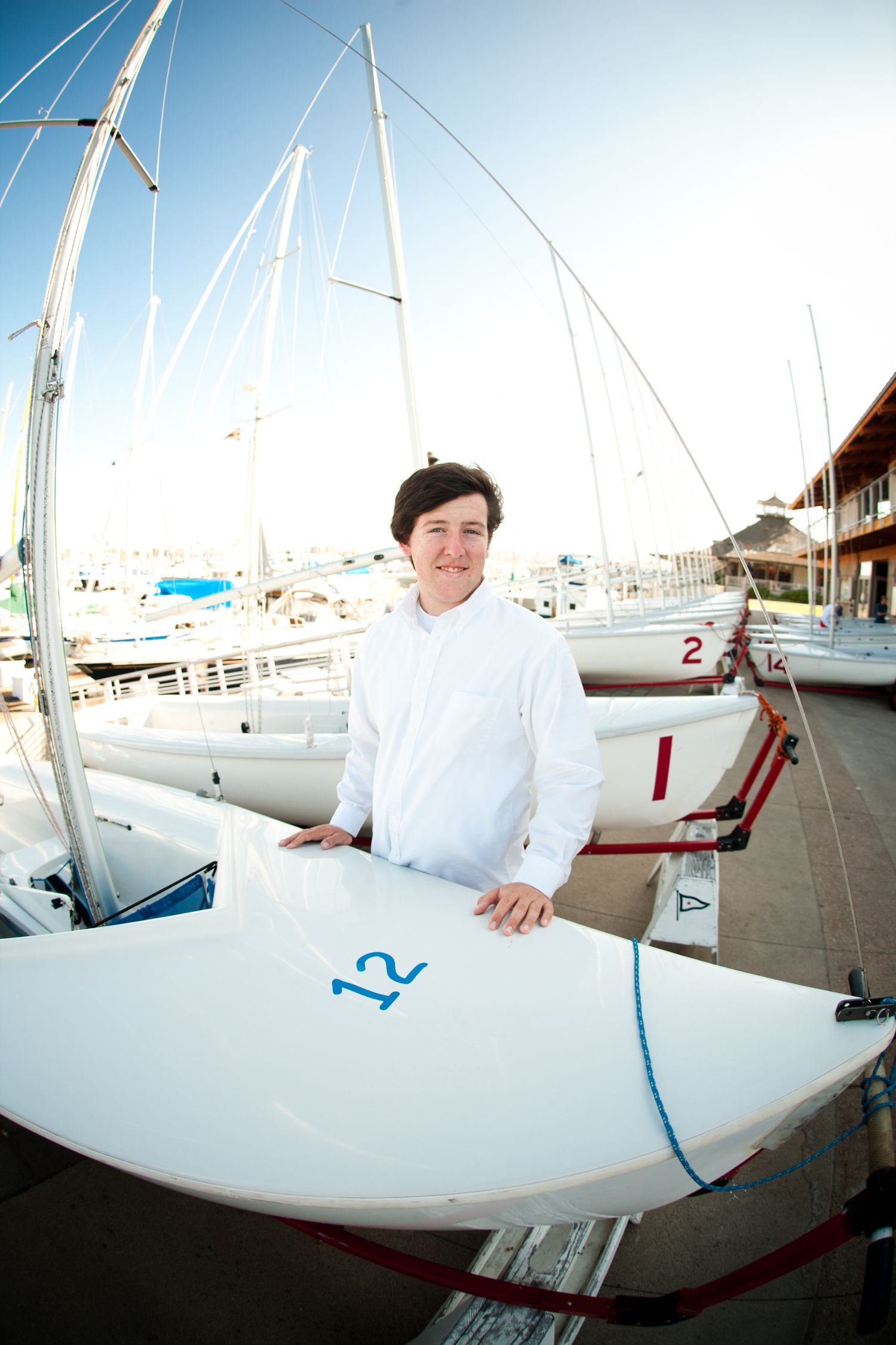 Person in white clothing stands next to a sailboat at a marina dock.