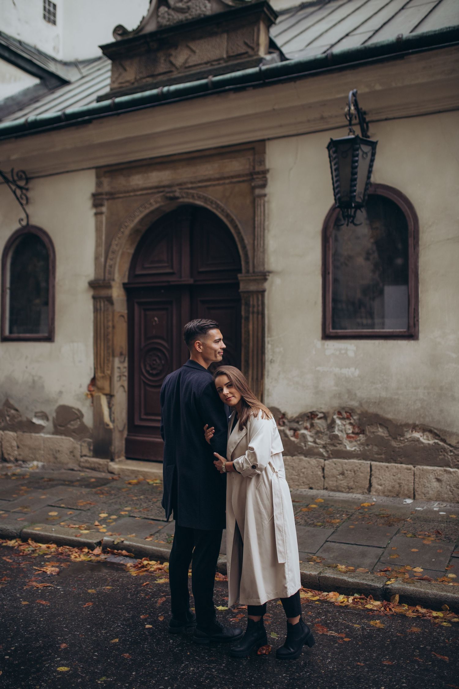 Couple embracing outside historic building with arched doorway and vintage lantern on autumn day.