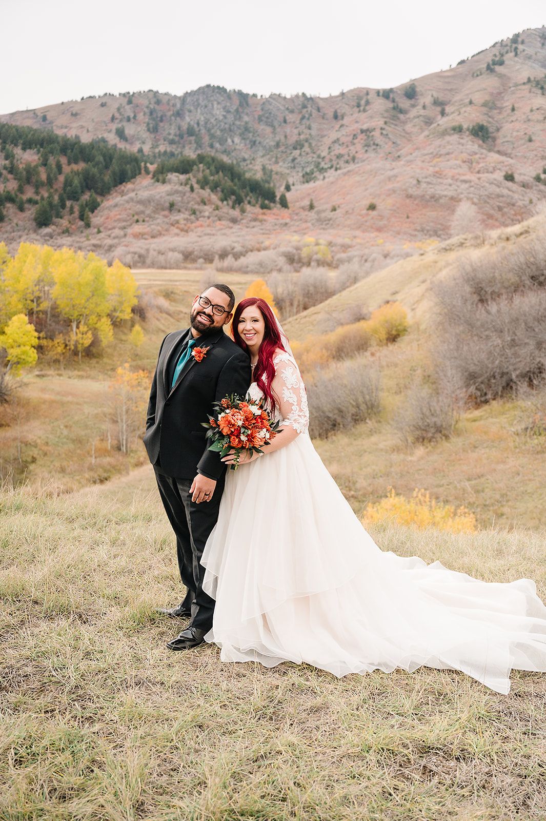 A wedding couple poses on a hillside with golden autumn foliage and mountains in the background.