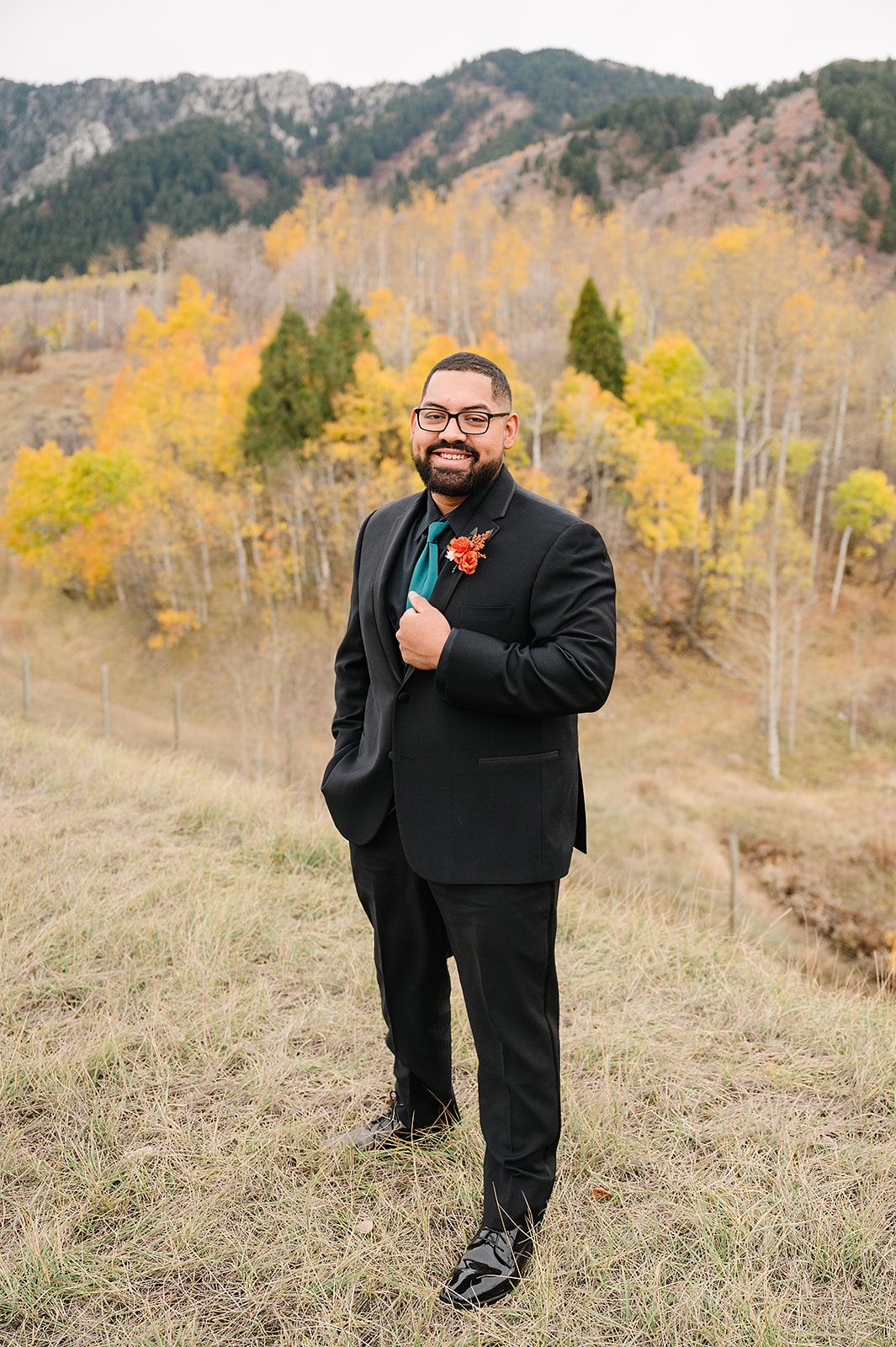 A groom in a classic black suit poses on a hillside with autumn colors in the background.