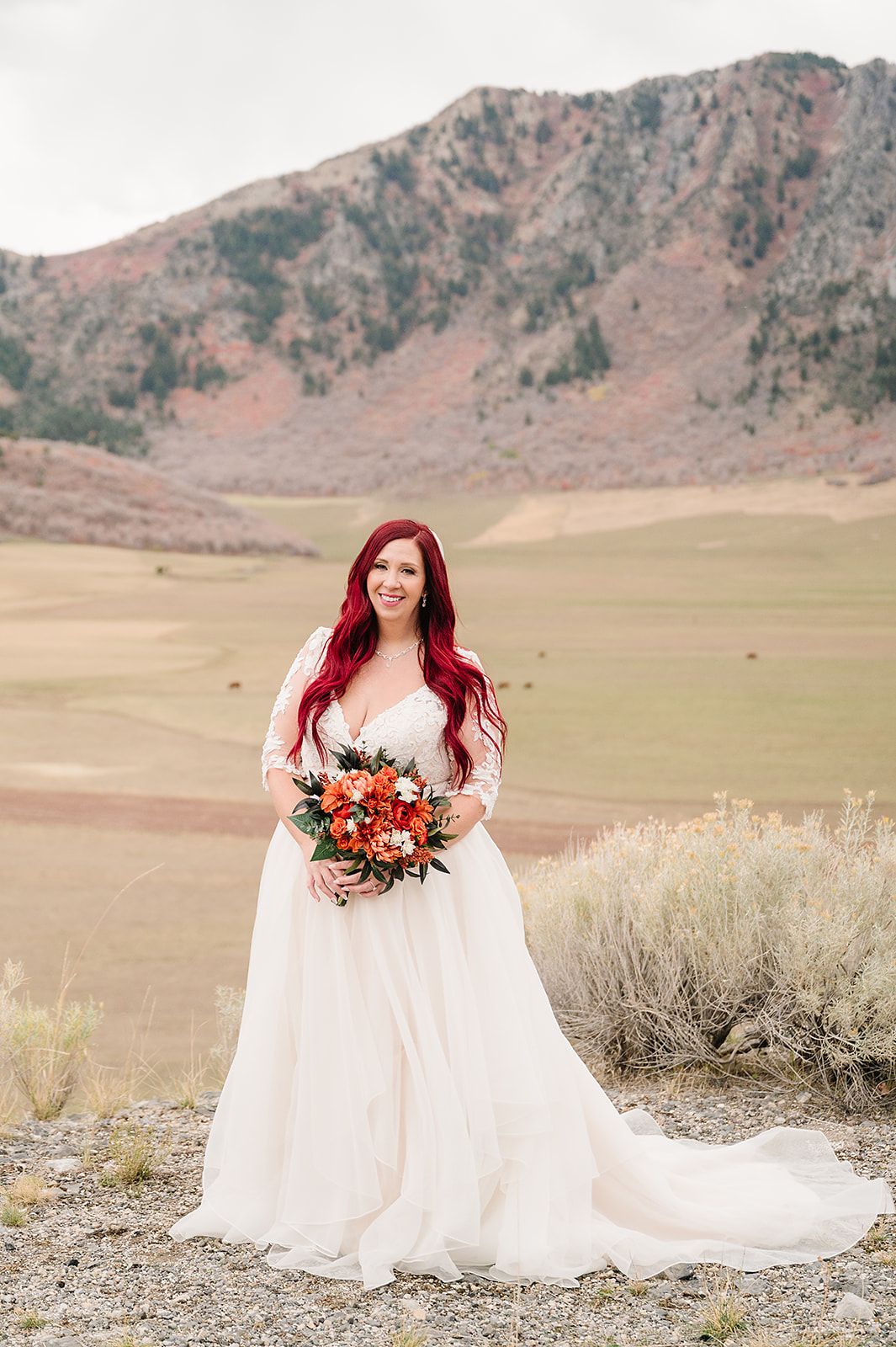A bride in a flowing white gown holds a vibrant fall bouquet against a mountainous backdrop.