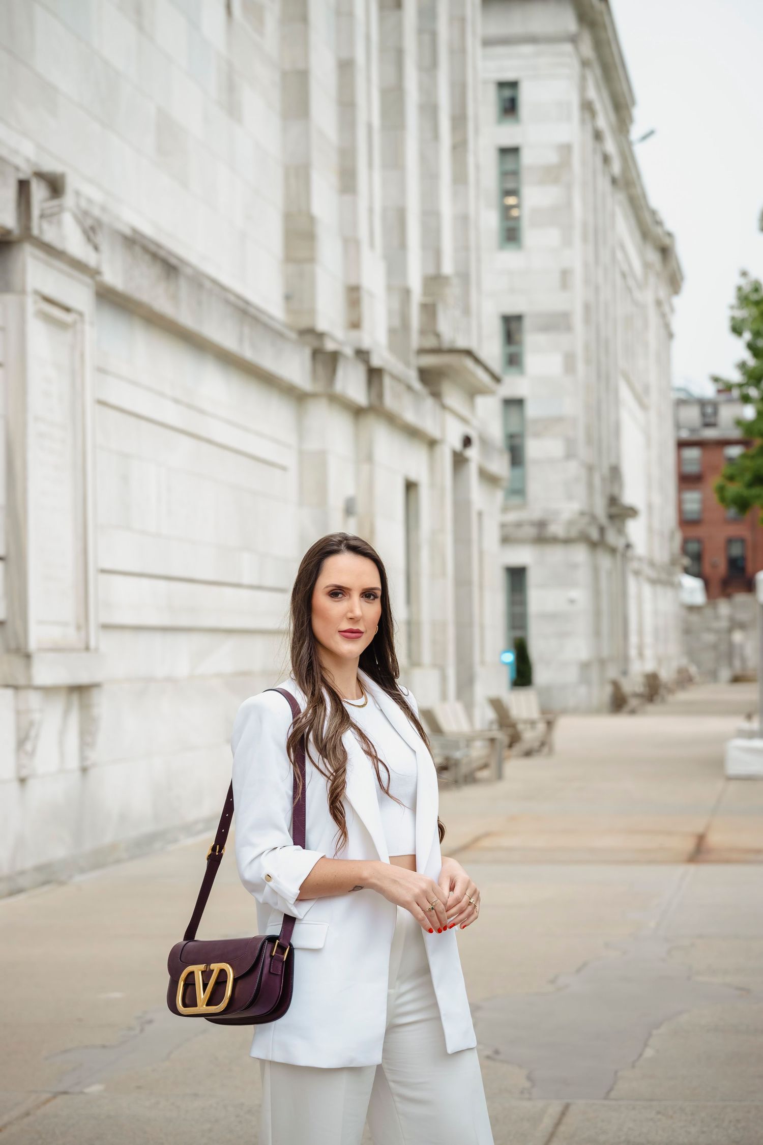 Stylish white pantsuit outfit accessorized with brown designer handbag against classic architectural backdrop.