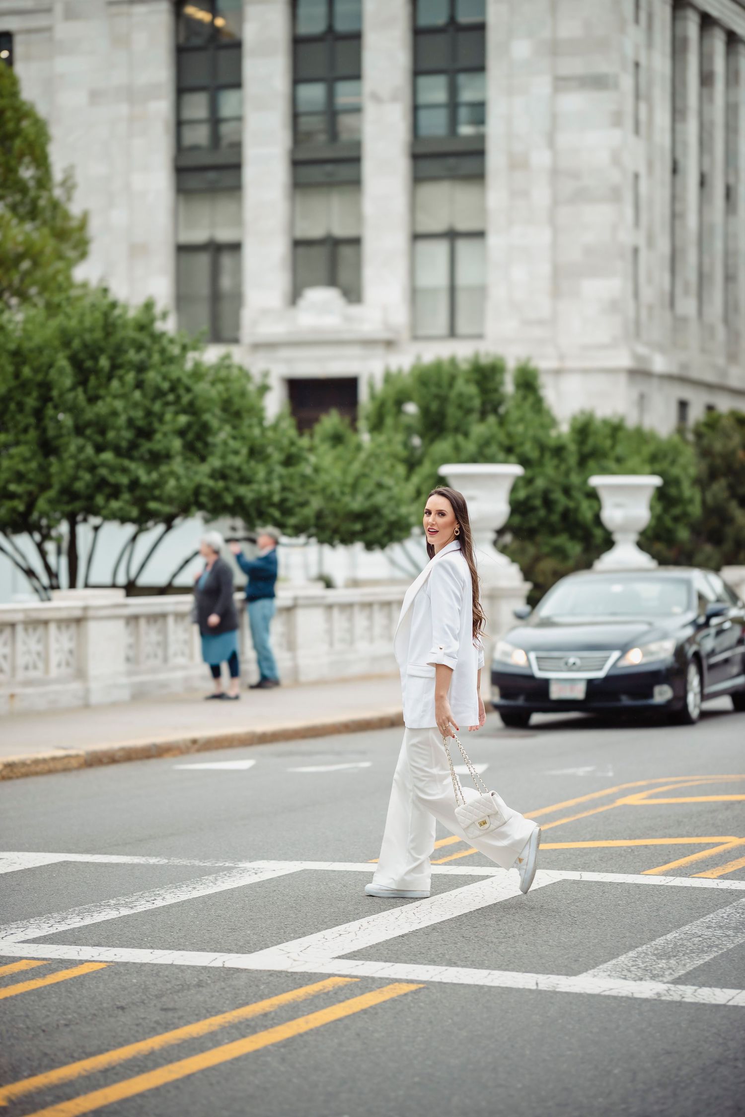 Person in all-white outfit crossing street in front of stone building with columns and landscaping.