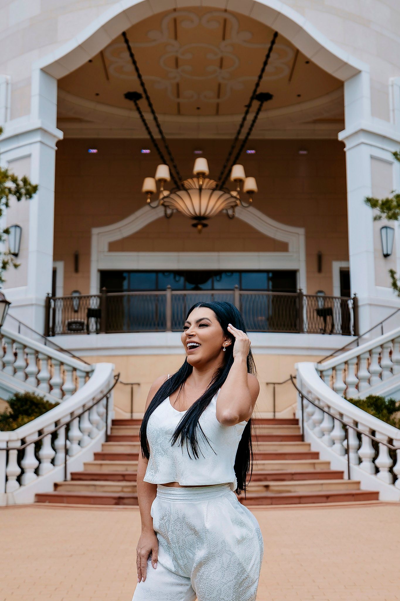 A two-piece white outfit photographed at an elegant entrance with grand staircases and decorative chandelier in the background.