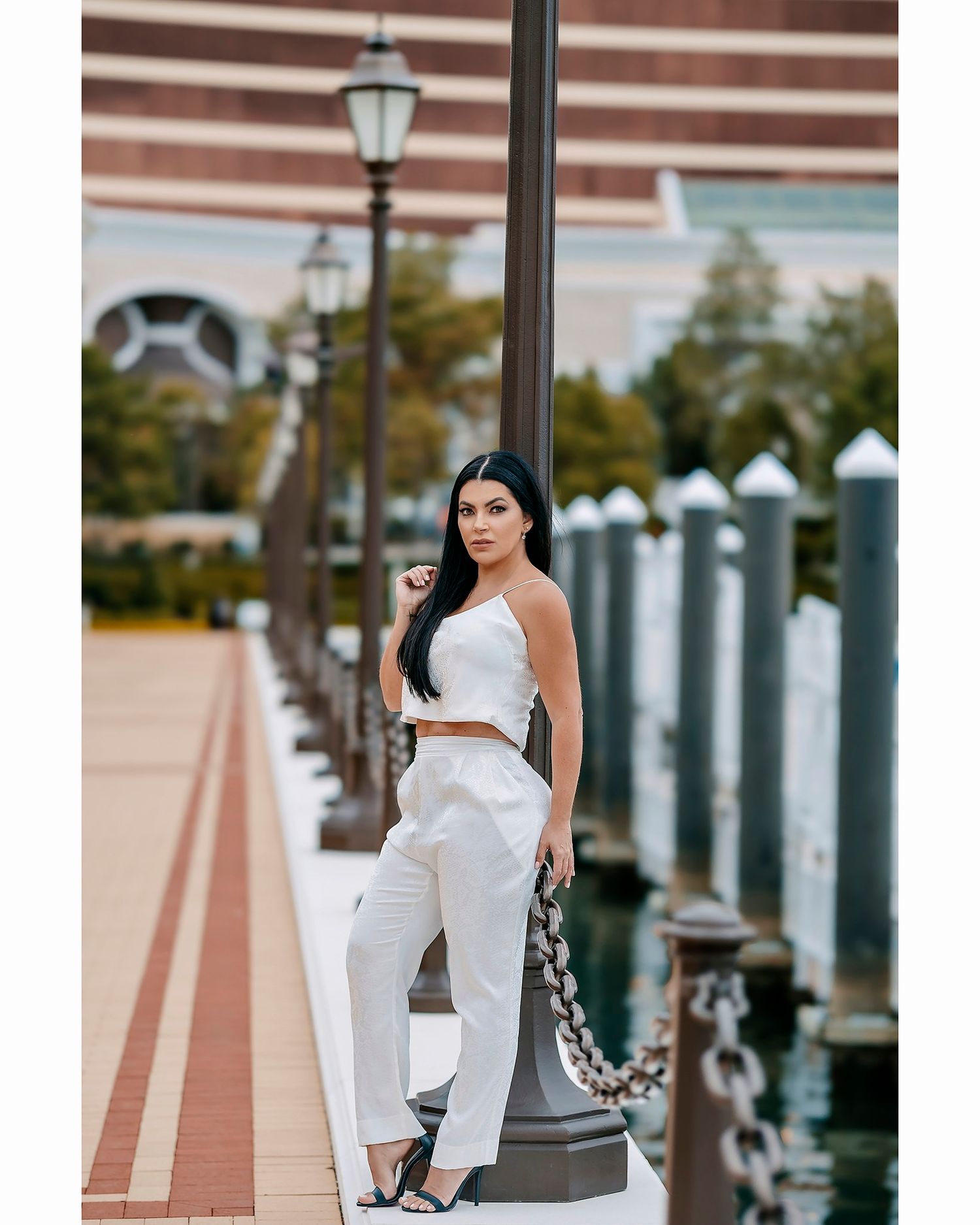 Woman in white outfit posing against waterfront railing with city buildings and streetlamp in background.
