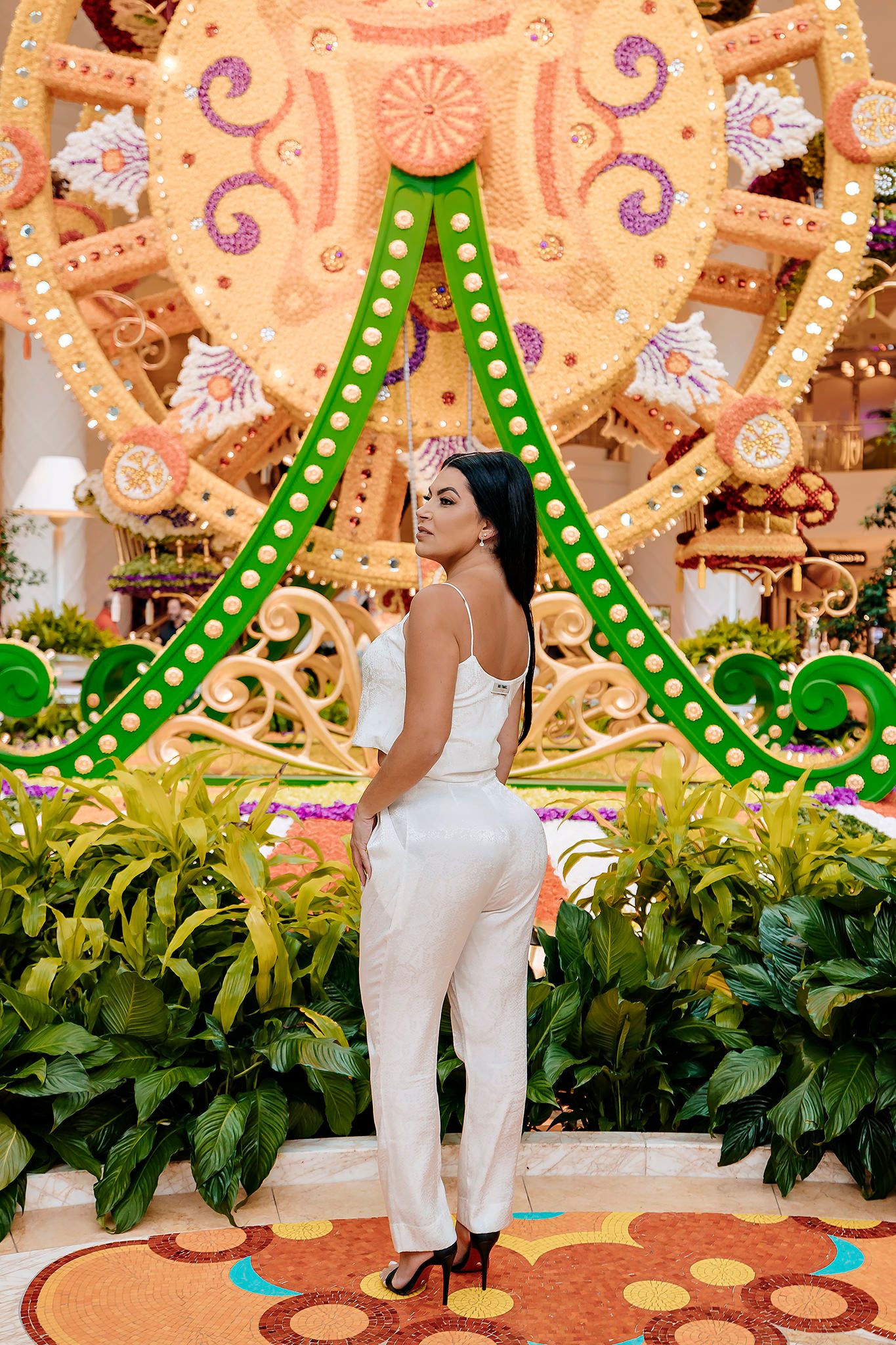 Elegant figure in white jumpsuit poses in front of decorative carnival backdrop with green lit archway and tropical plants.