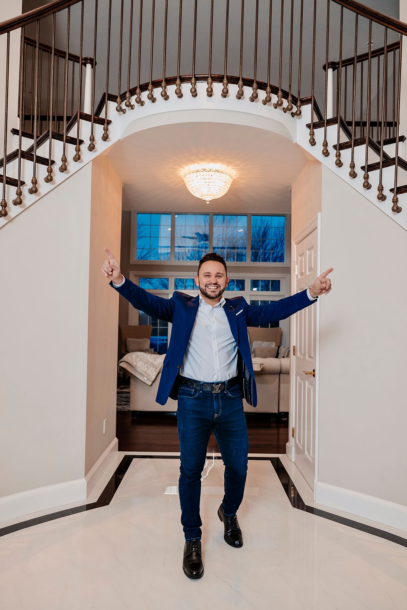 Person celebrating with arms raised under elegant staircase with chandelier in luxury home entrance.