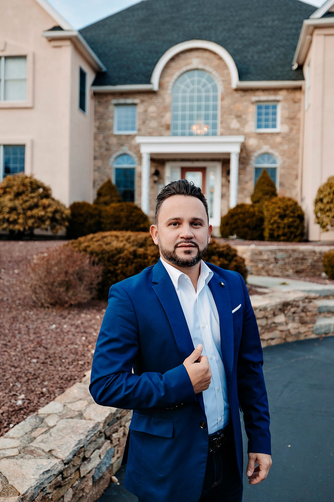 Person in a navy blue suit jacket stands outside a luxurious stone and brick house with arched windows.