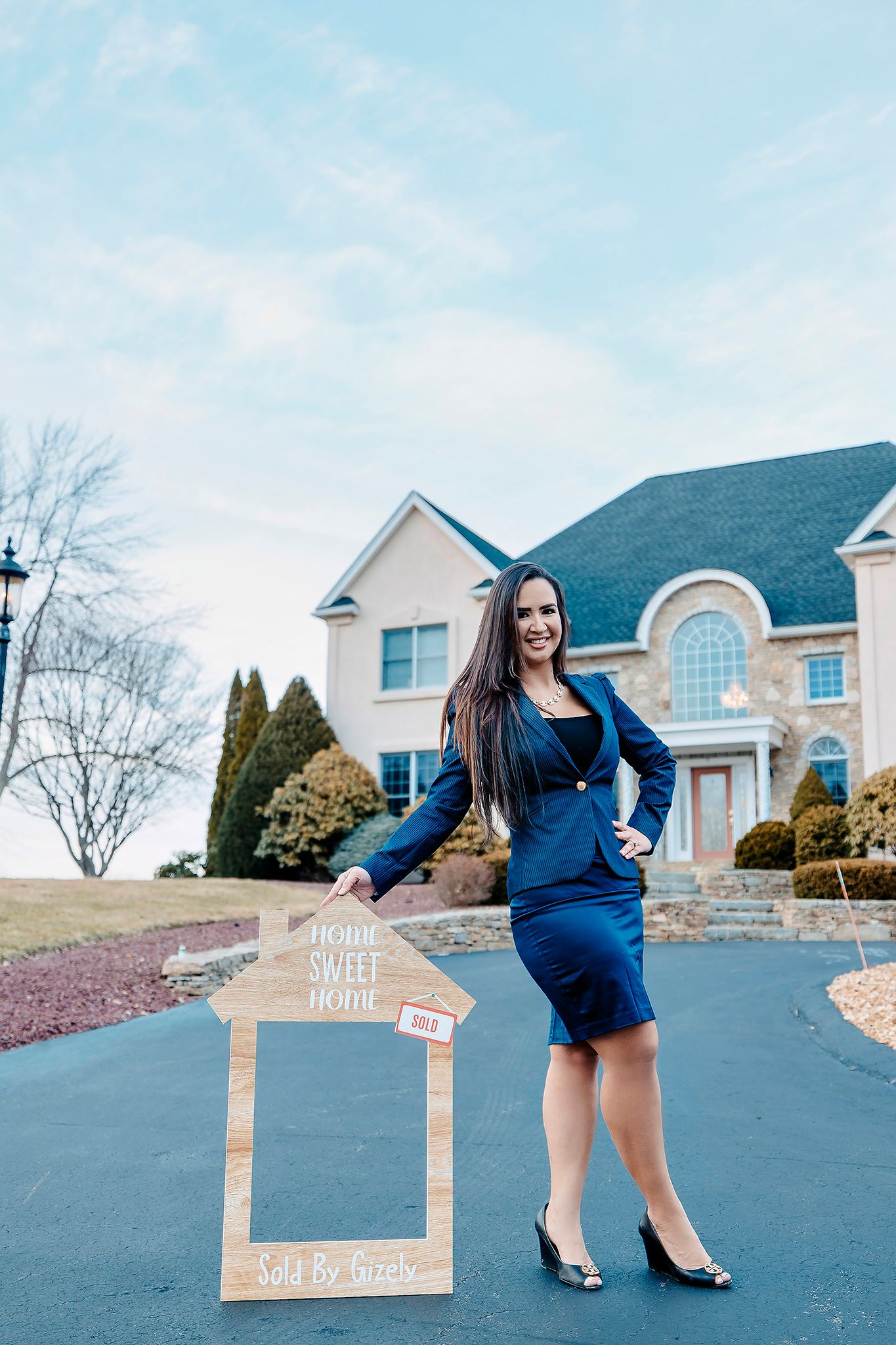 Professional wearing navy blue business attire poses with a decorative frame in front of a luxury home.