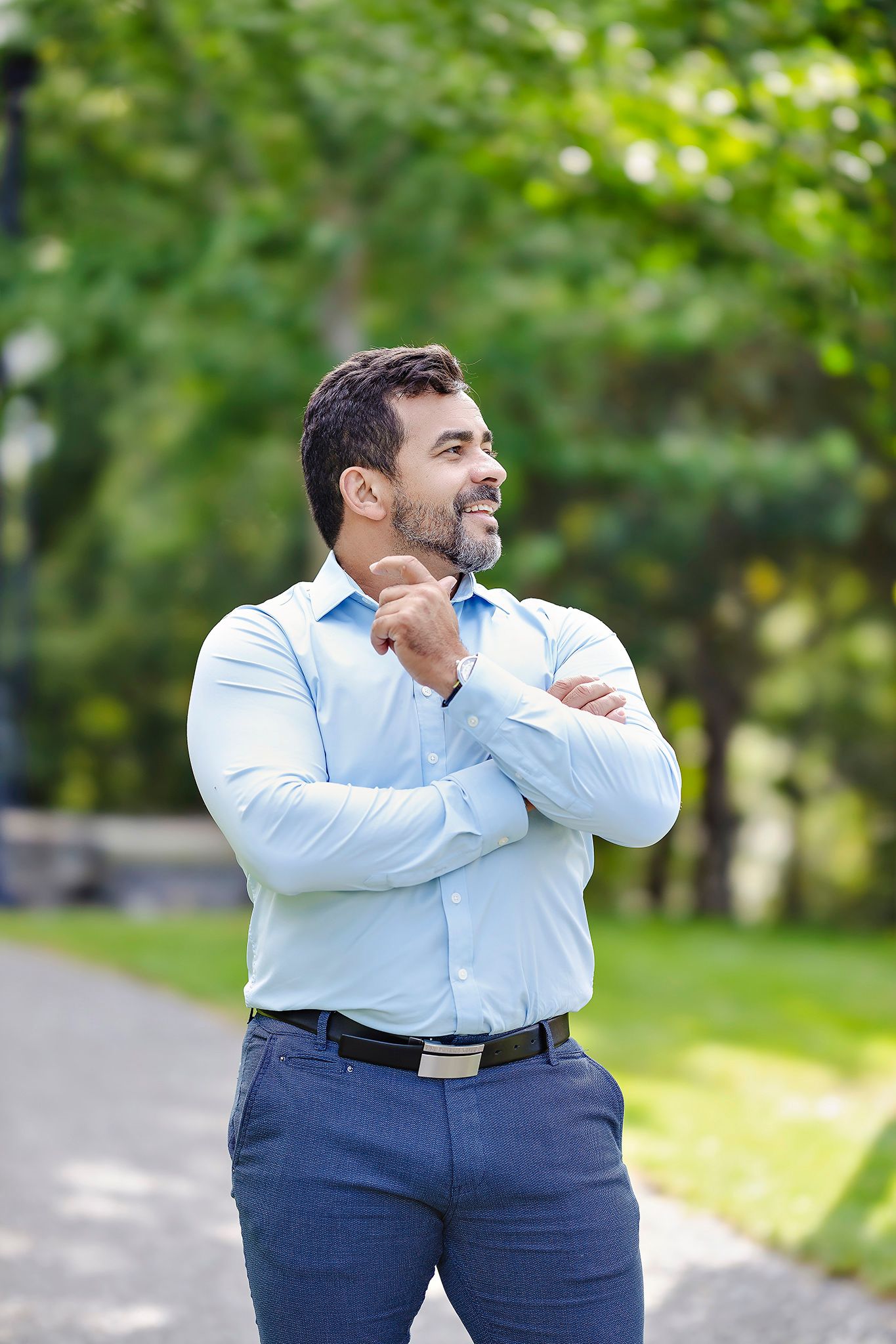 A man in a light blue dress shirt and navy pants poses outdoors in a park setting.