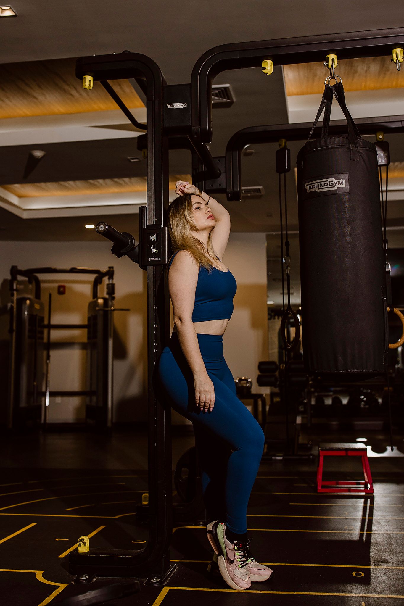 Athletic figure in blue workout set poses by punching bag in modern gym setting.
